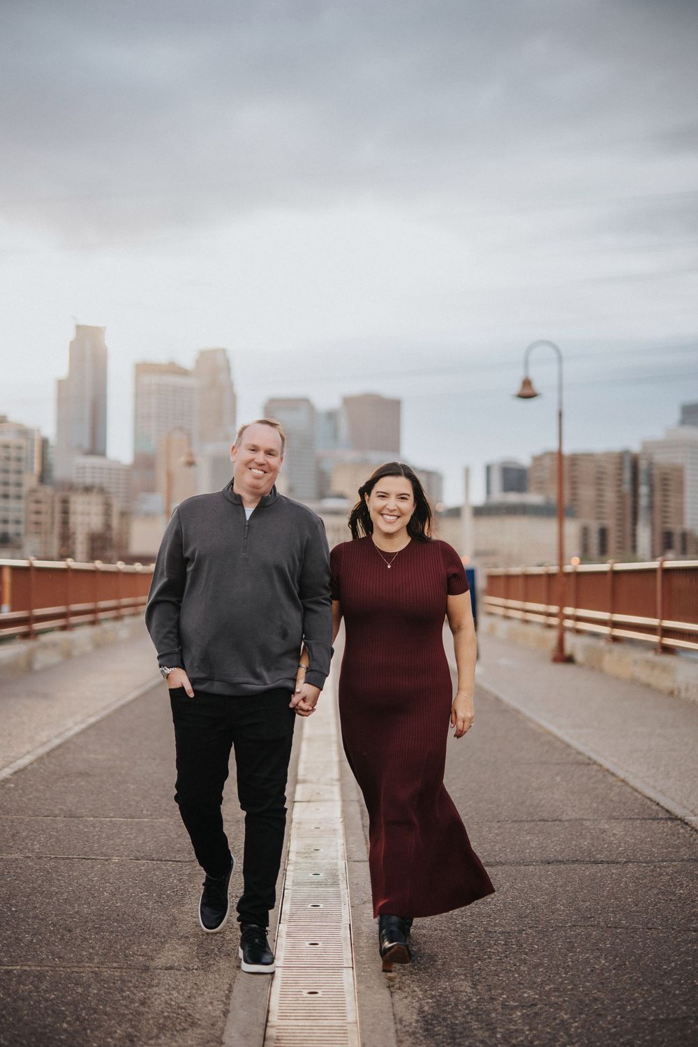 Couple walking and laughing together on an urban bridge with Minneapolis skyline in the background on cloudy day.