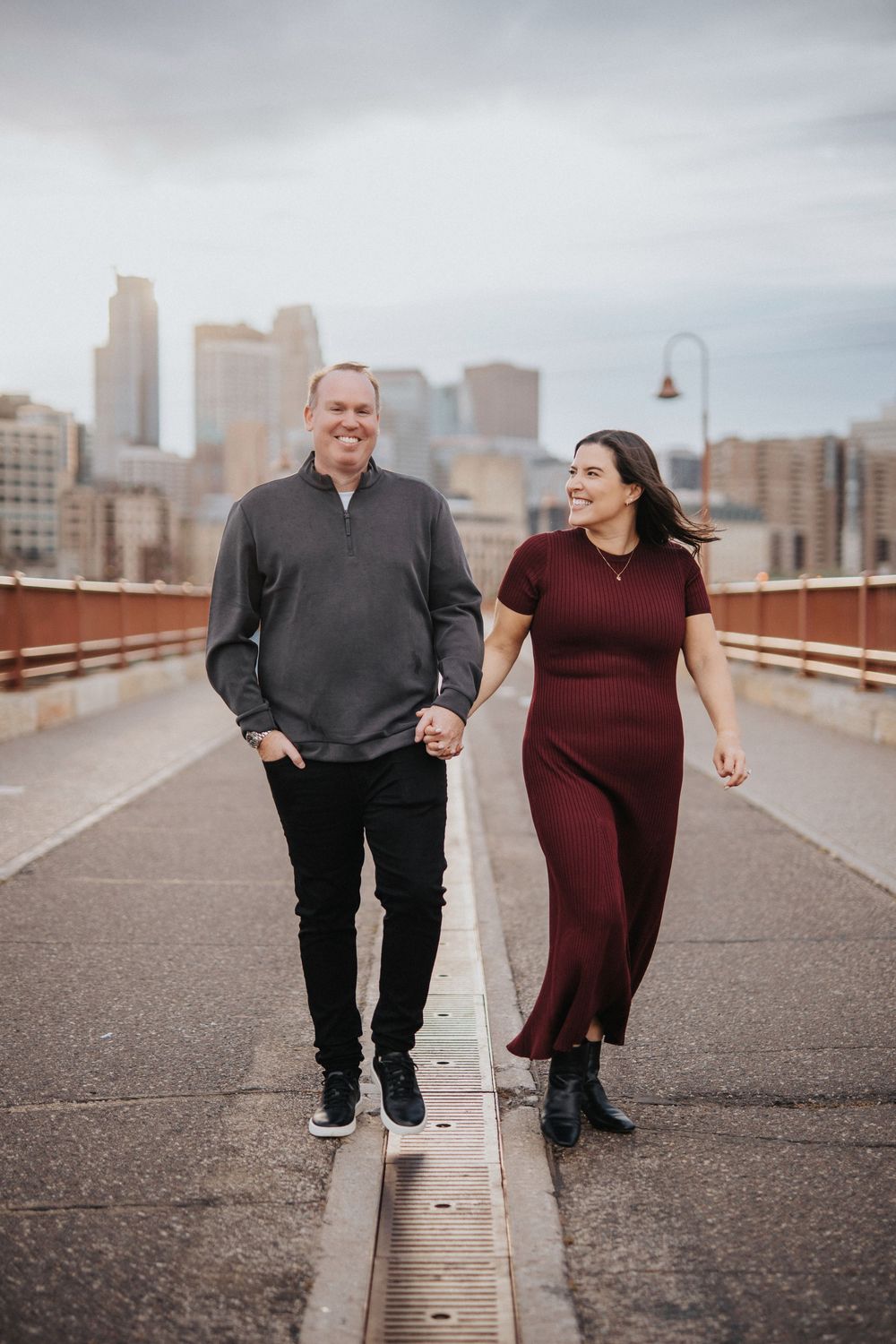 A couple in burgundy and black outfits walks playfully along an urban pathway with city buildings in background.