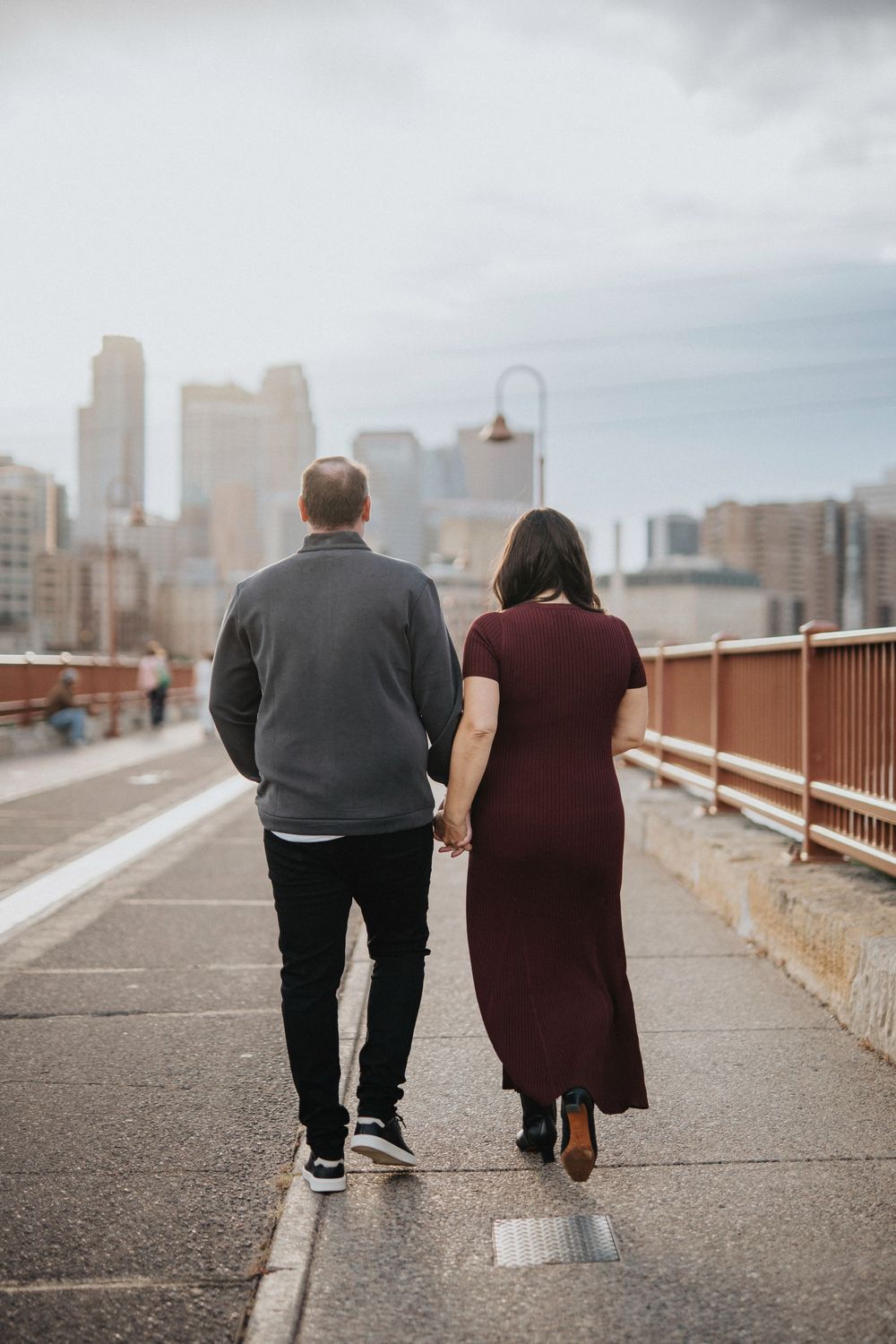 A couple walks together on a concrete bridge pathway with urban buildings visible in the distance.
