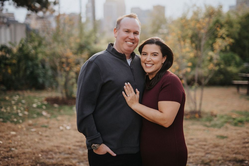 A couple embraces outdoors during an autumn engagement photo session with fall foliage in the background.