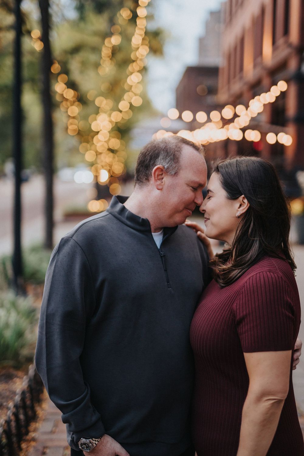 Romantic couple embracing near twinkling lights and brick buildings at dusk.