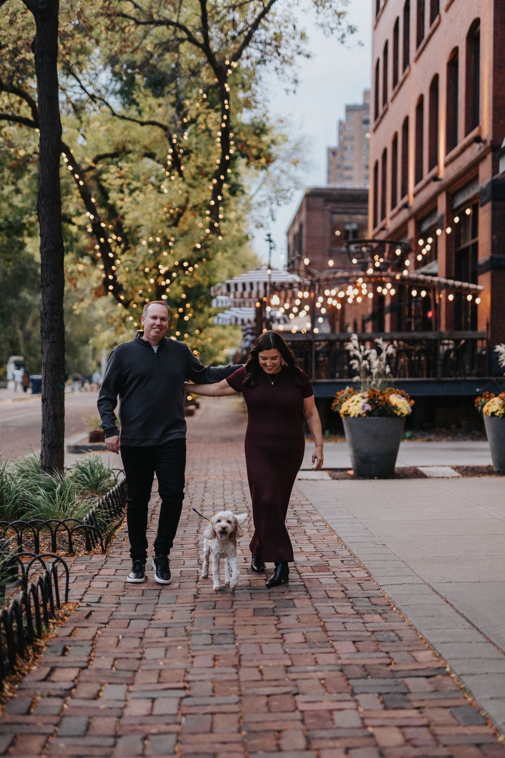 A couple walks their small white dog along a brick sidewalk with string lights overhead in an urban setting.