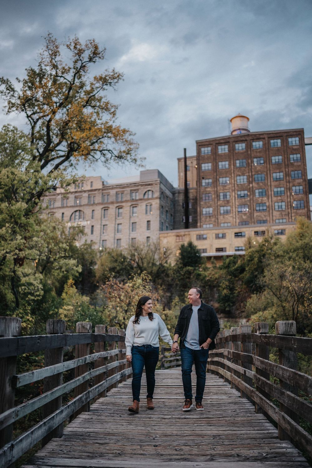 A couple walks together on a wooden bridge pathway surrounded by autumn trees and brick buildings on a cloudy day.