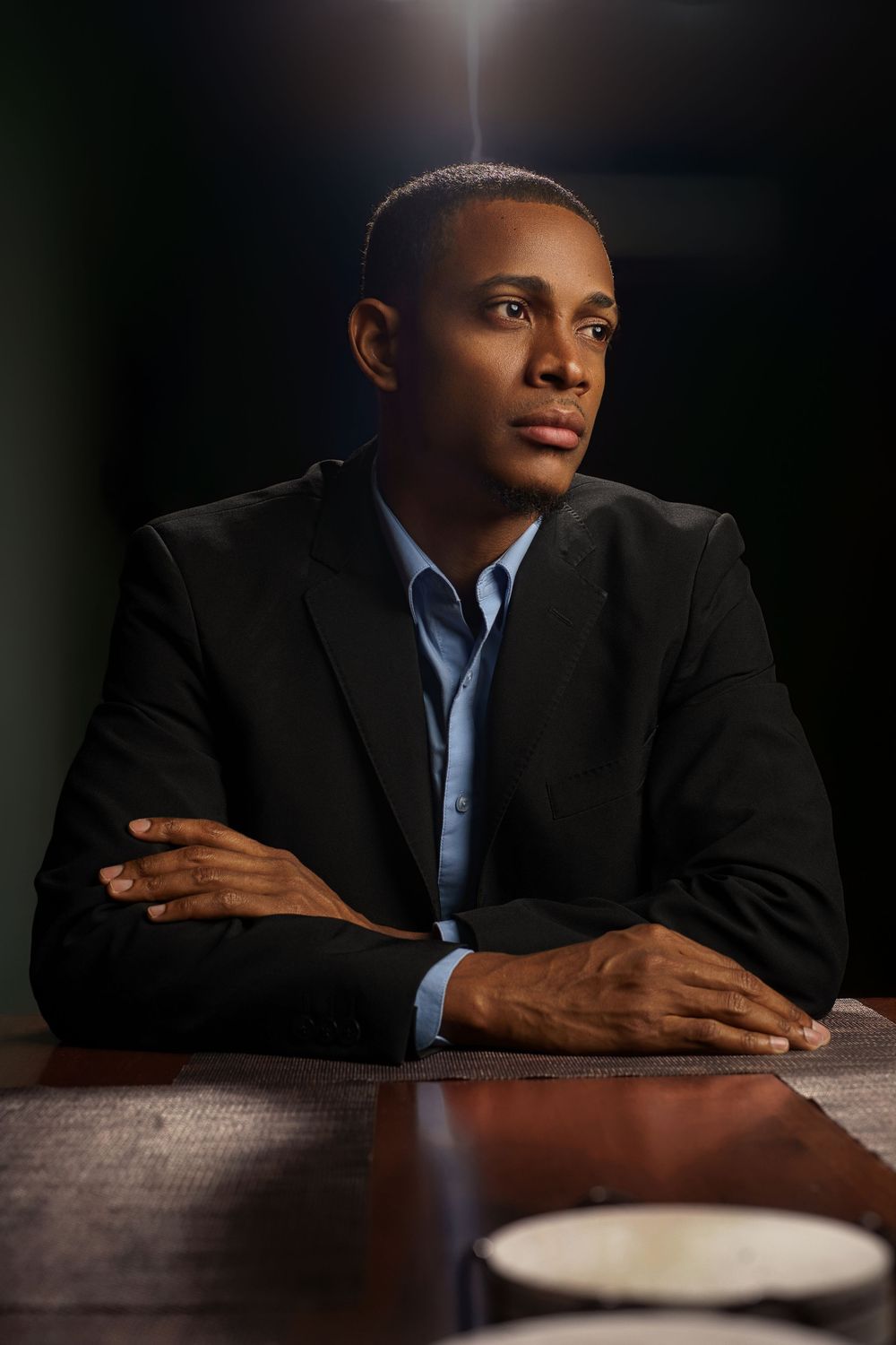 A person in a dark business suit and blue shirt sits at a wooden table in dramatic low-key lighting.