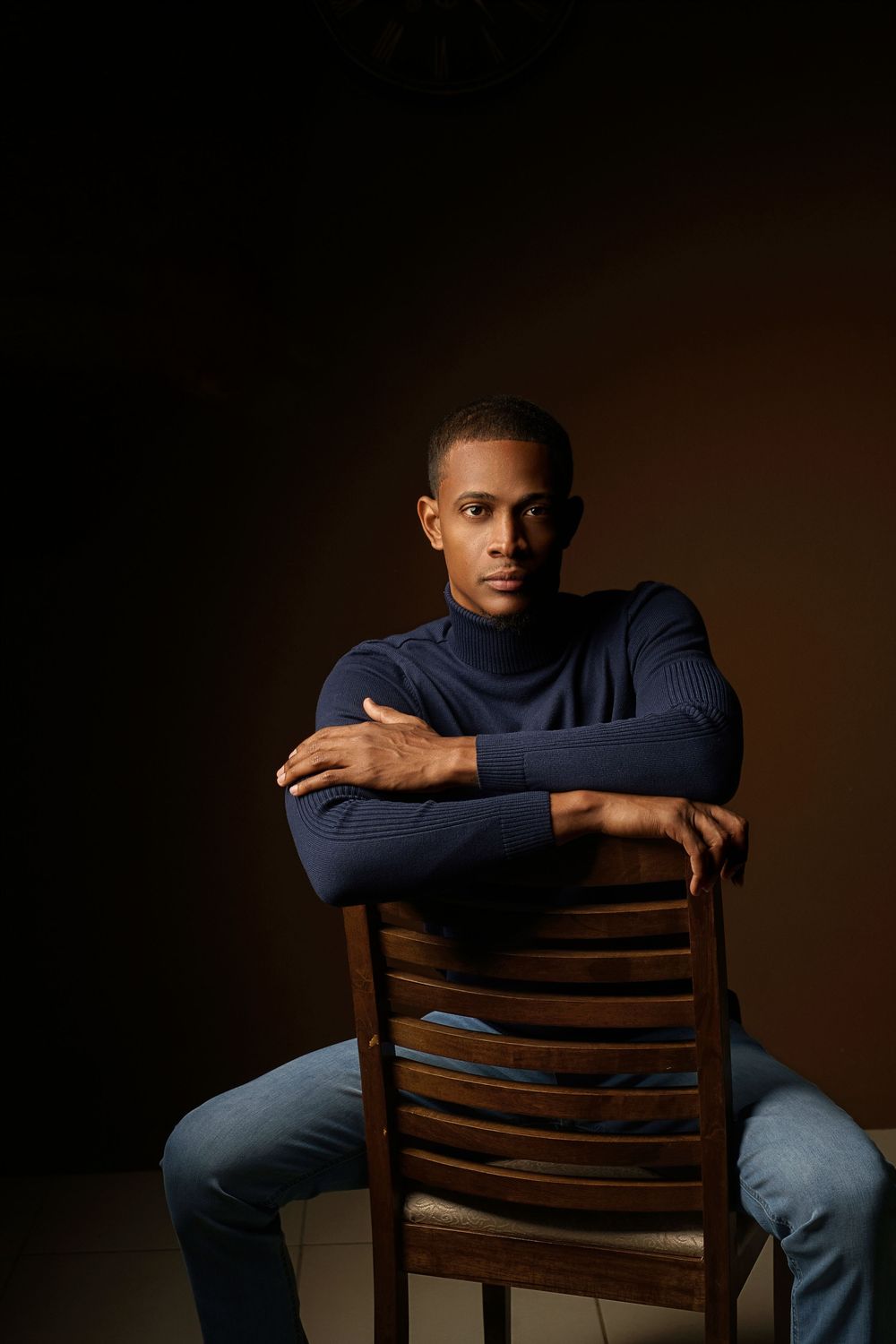 Dramatic studio portrait of person in navy sweater sitting backwards on wooden chair against dark background.