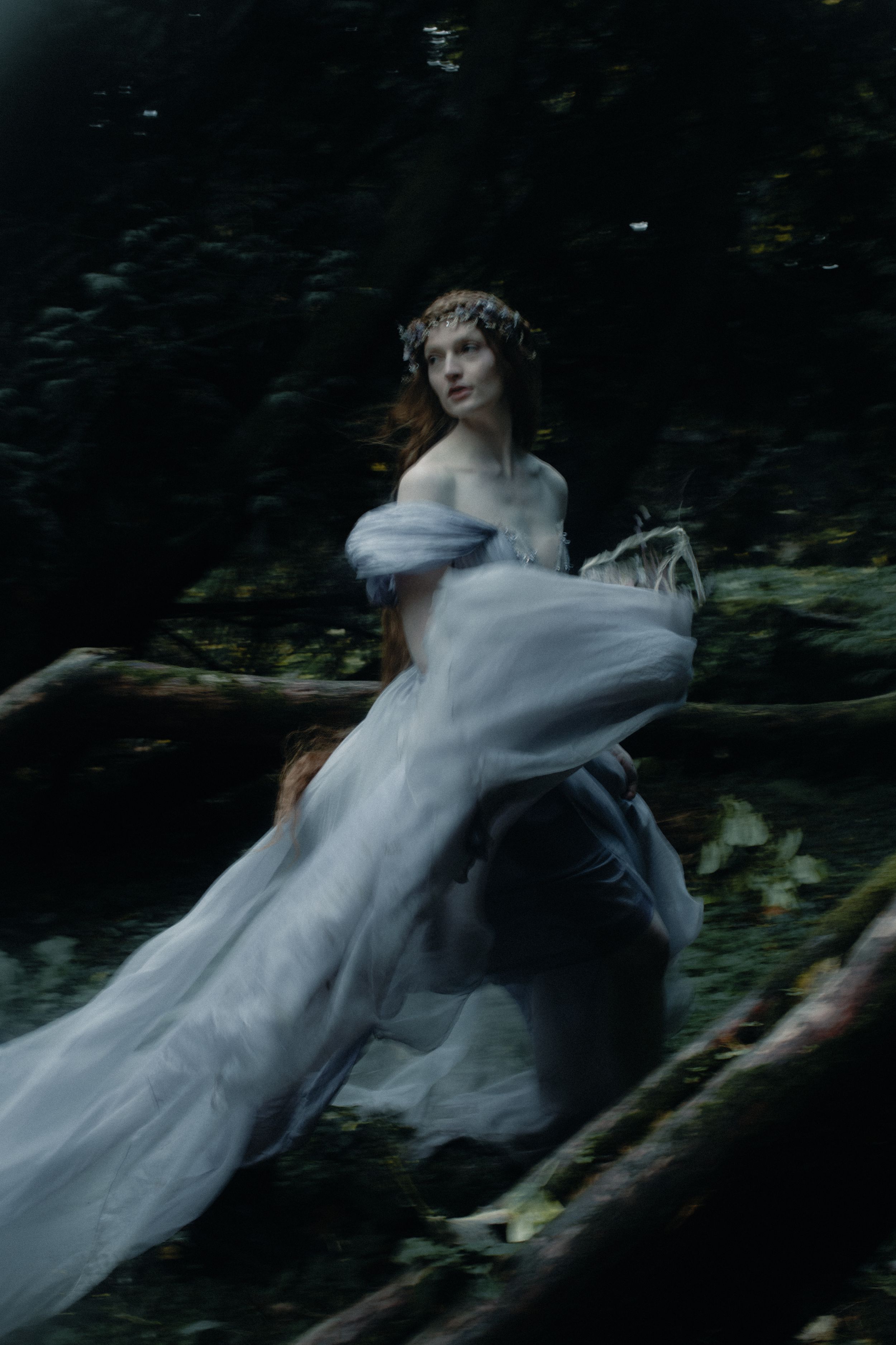 A figure in a flowing white dress and floral crown runs through a dark forest in an ethereal, dreamlike scene.