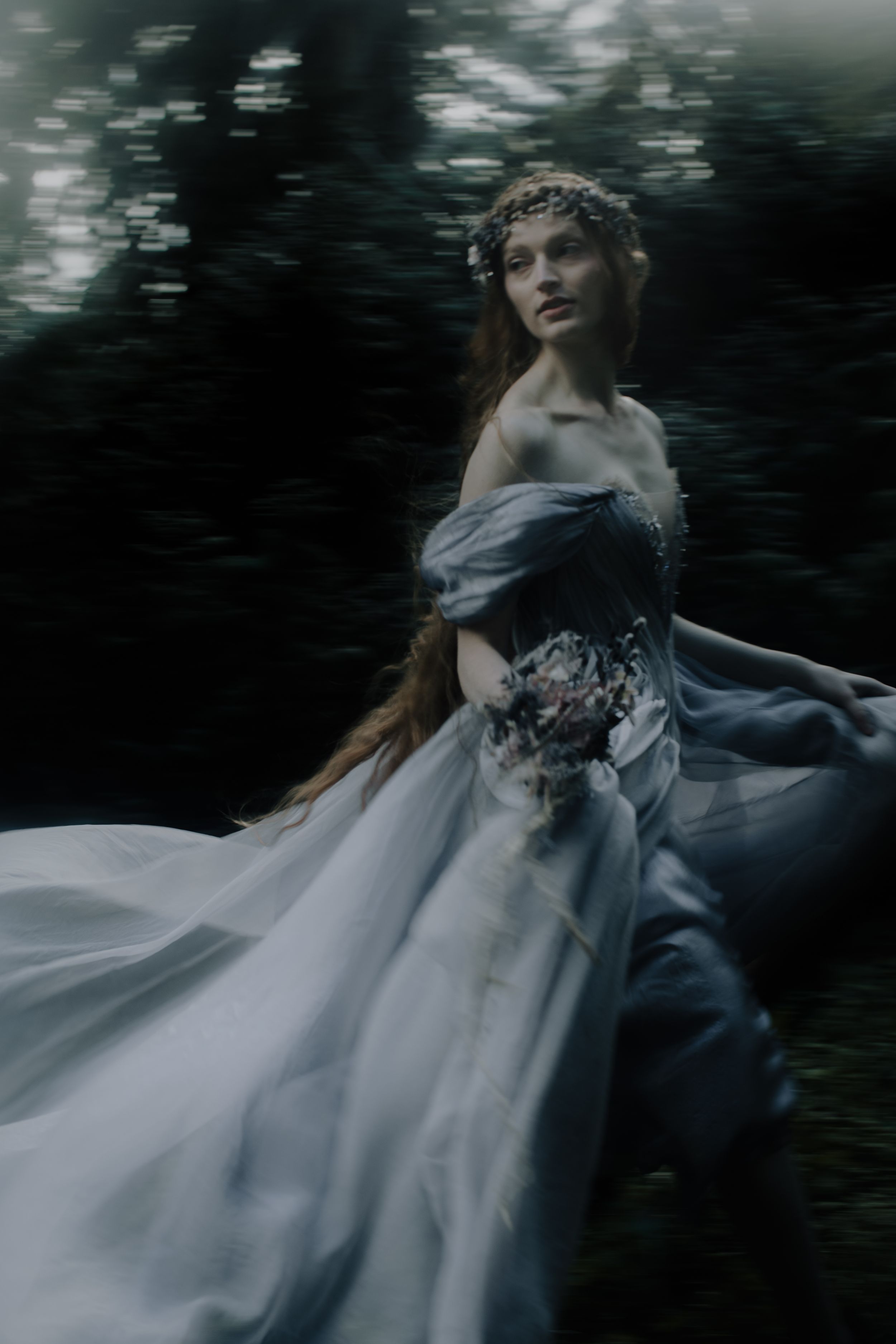 A figure in a flowing blue and white wedding gown twirls dramatically against a dark forest backdrop in motion blur.