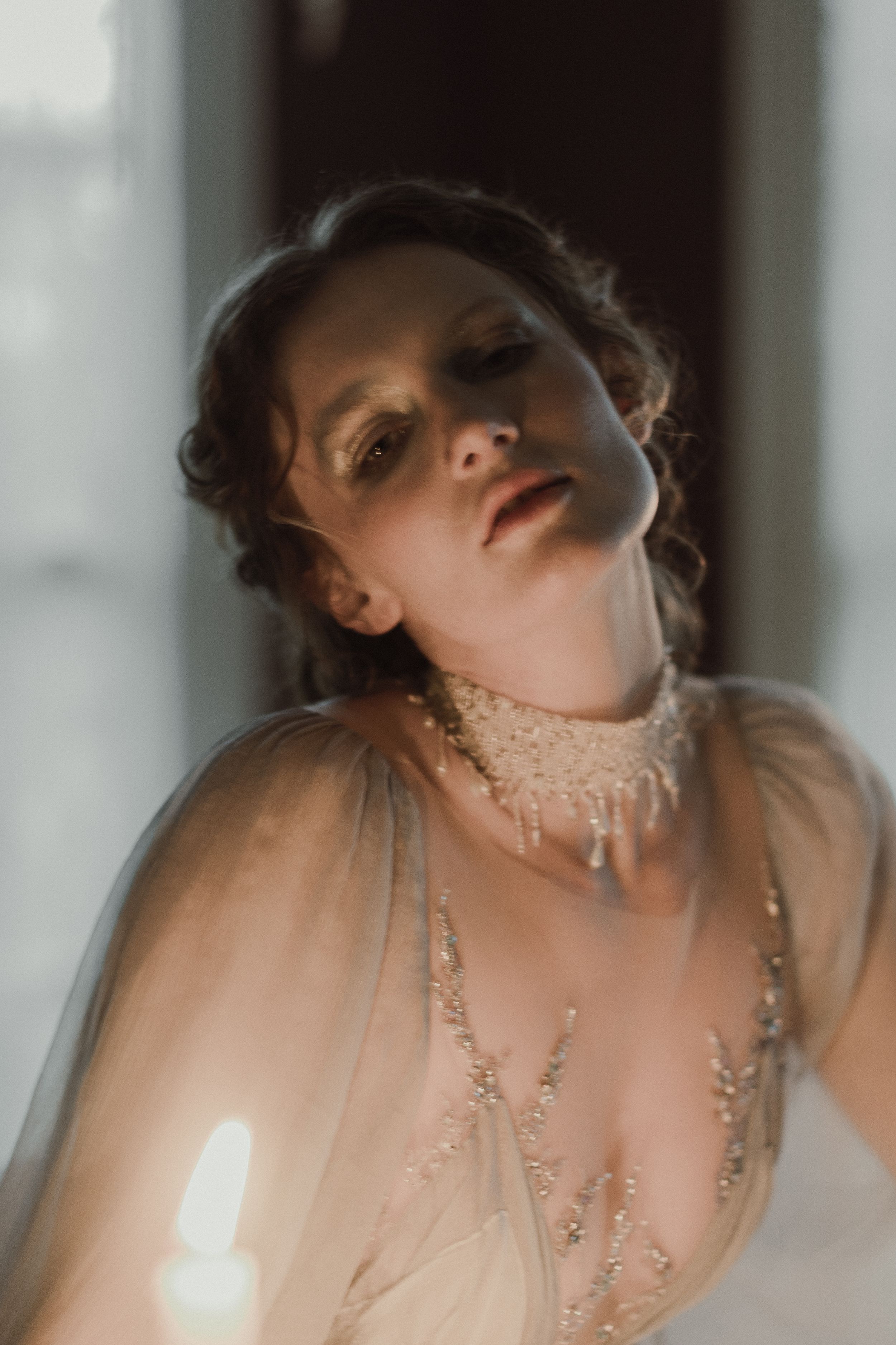 Artistic boudoir photo with beaded neckpiece and sheer fabric in soft window light.