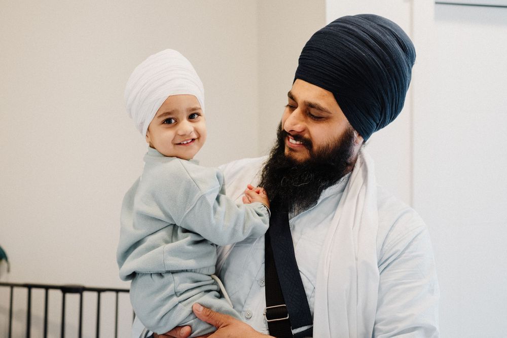 Sikh father with dark beard and blue turban holding smiling toddler in white turban.