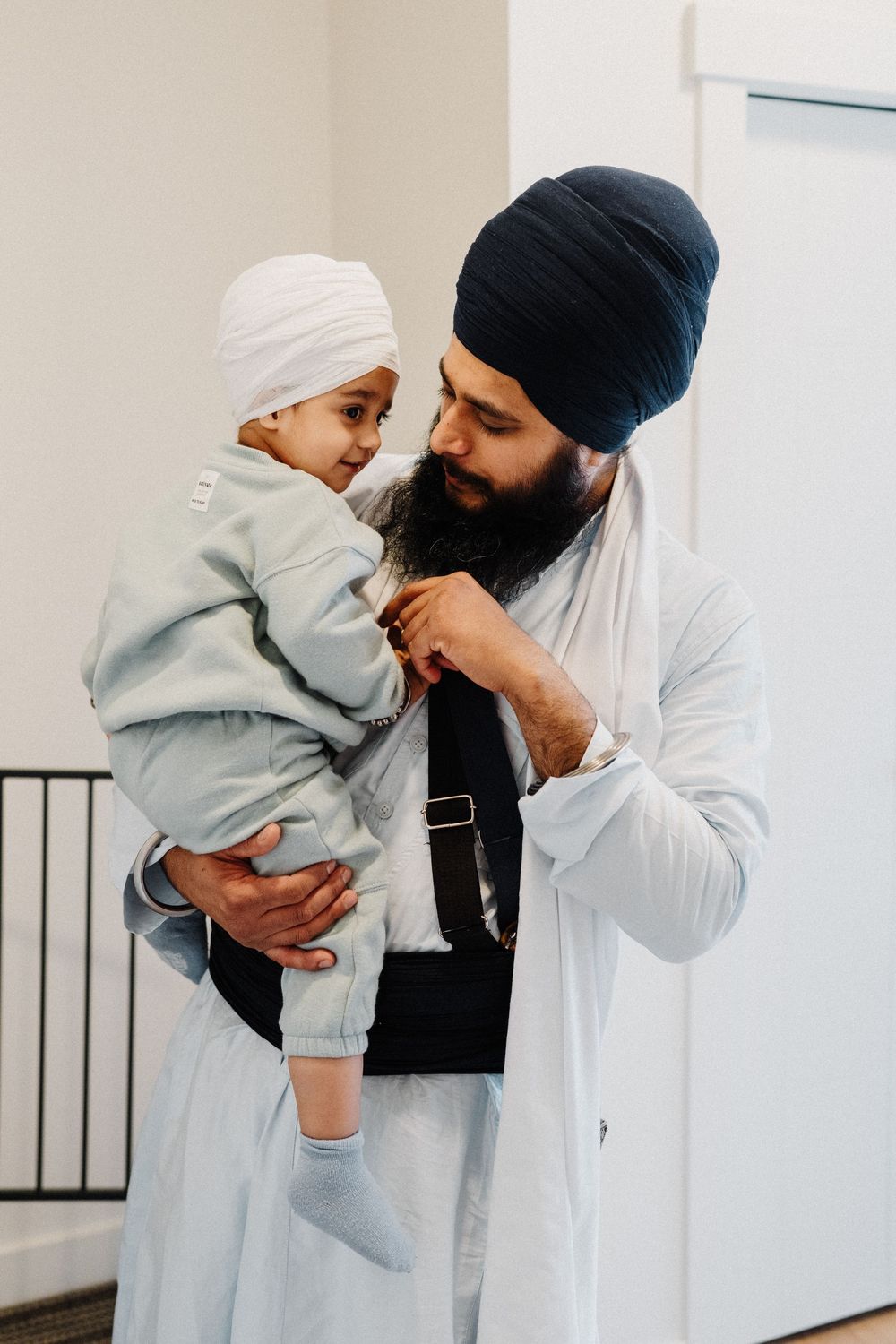 Bearded Sikh man in blue turban tenderly holding young child in white turban indoors.