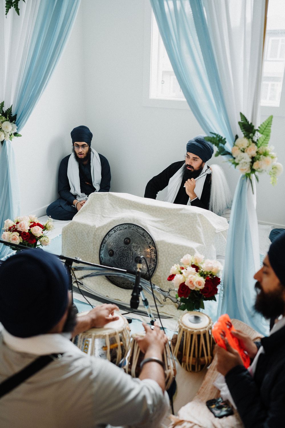 Sikh musicians playing tabla drums during a religious ceremony with blue drape decor.