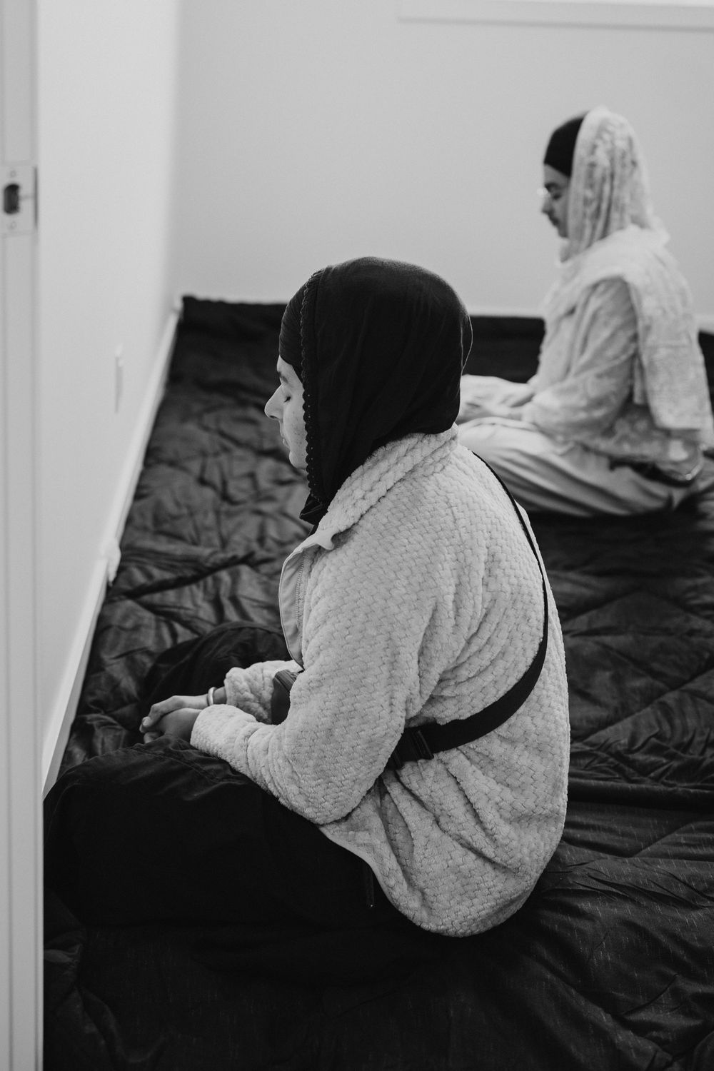 Two women in headscarves sitting quietly on black floor covering in black and white photo.