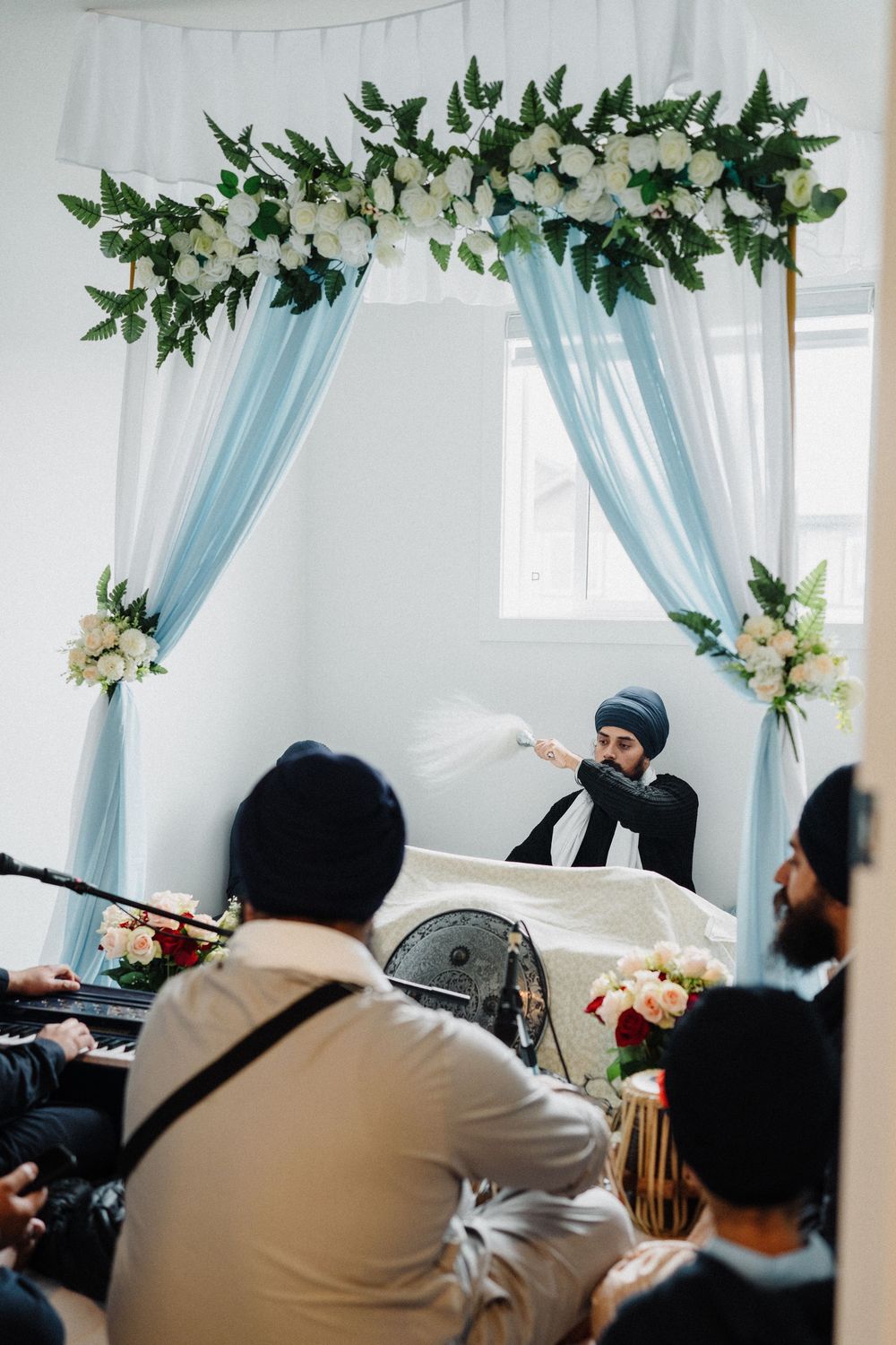 Sikh priest waving chaur sahib over holy scripture under floral arch with blue drapes.