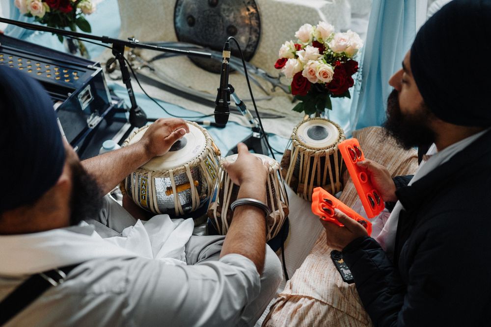 Two Sikh musicians playing tabla drums at a traditional wedding ceremony with flowers in background.