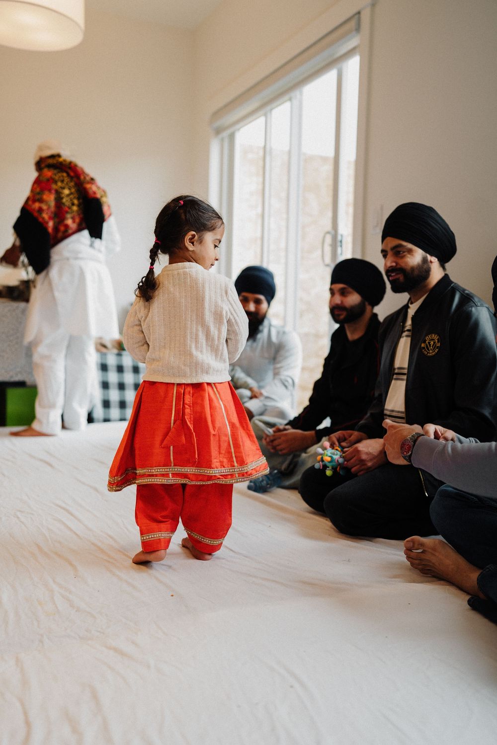 Young girl in red traditional Punjabi outfit dancing before seated Sikh men wearing turbans indoors.