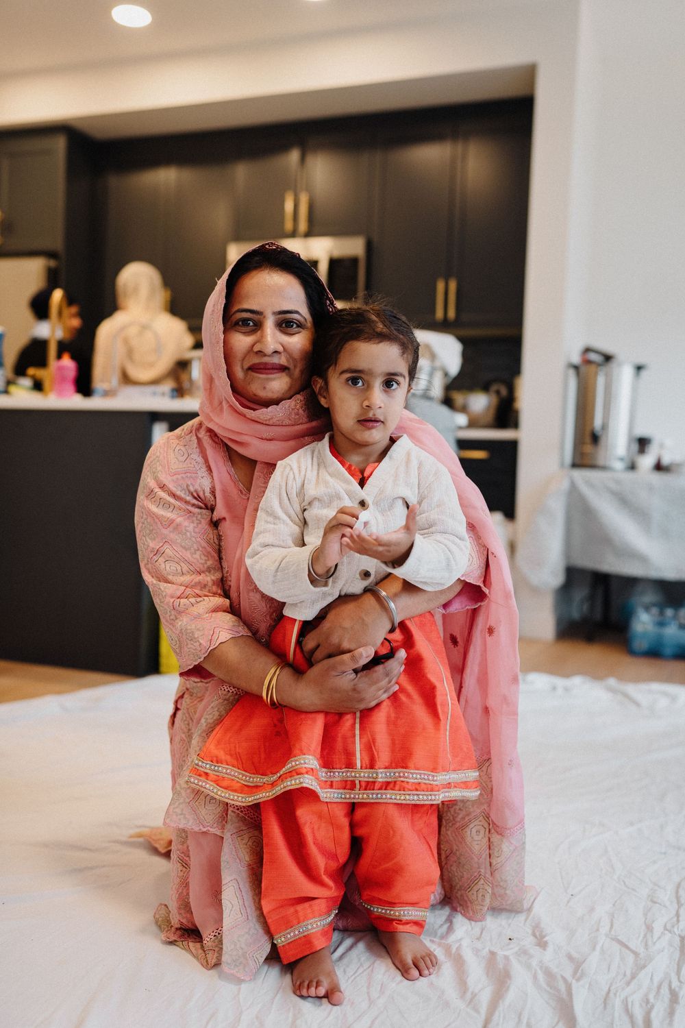 Woman in pink salwar kameez holding a young child in orange lehenga, posing together in a modern kitchen.