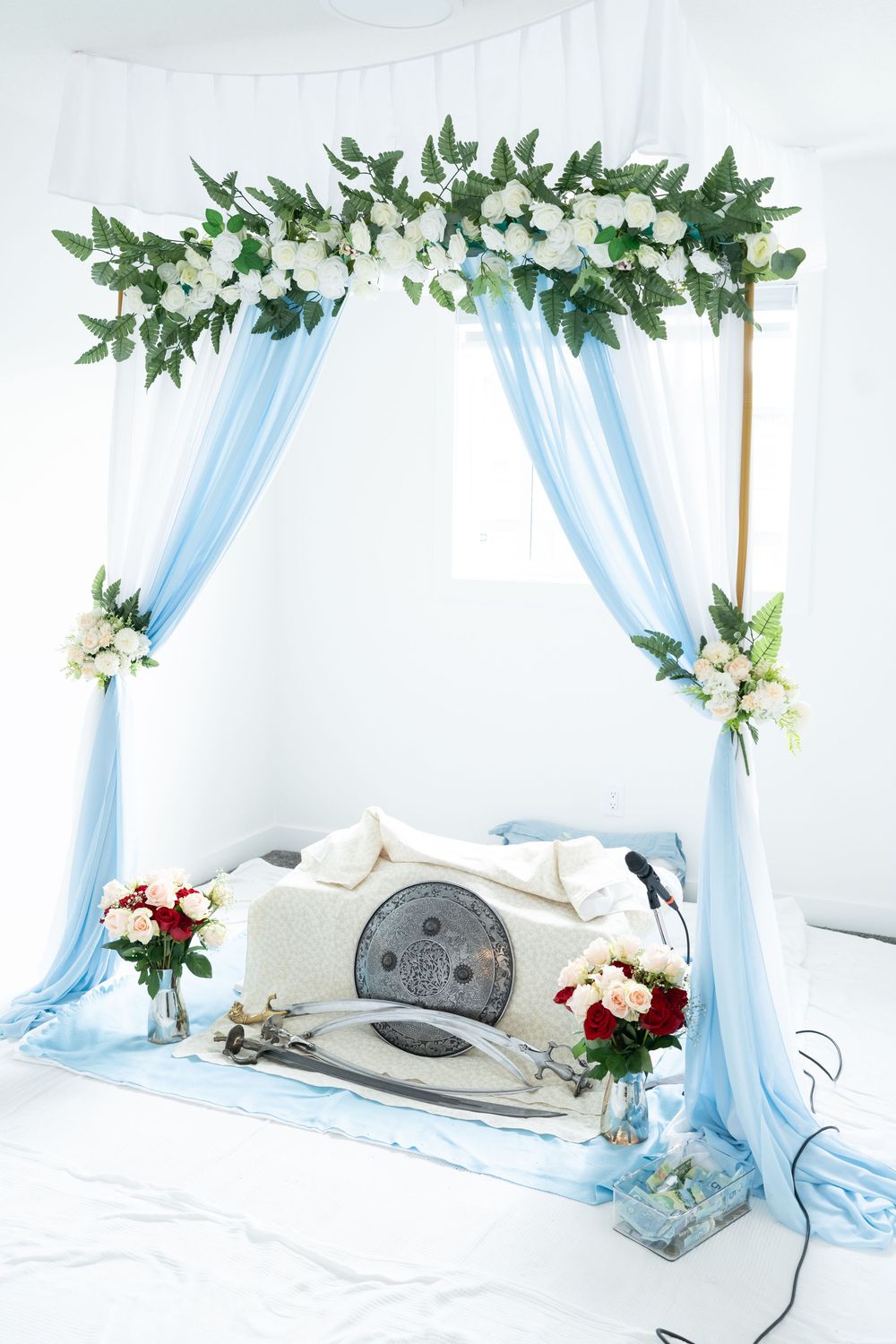 Blue draped wedding chuppah decorated with white flowers and greenery, with ceremonial items displayed beneath.