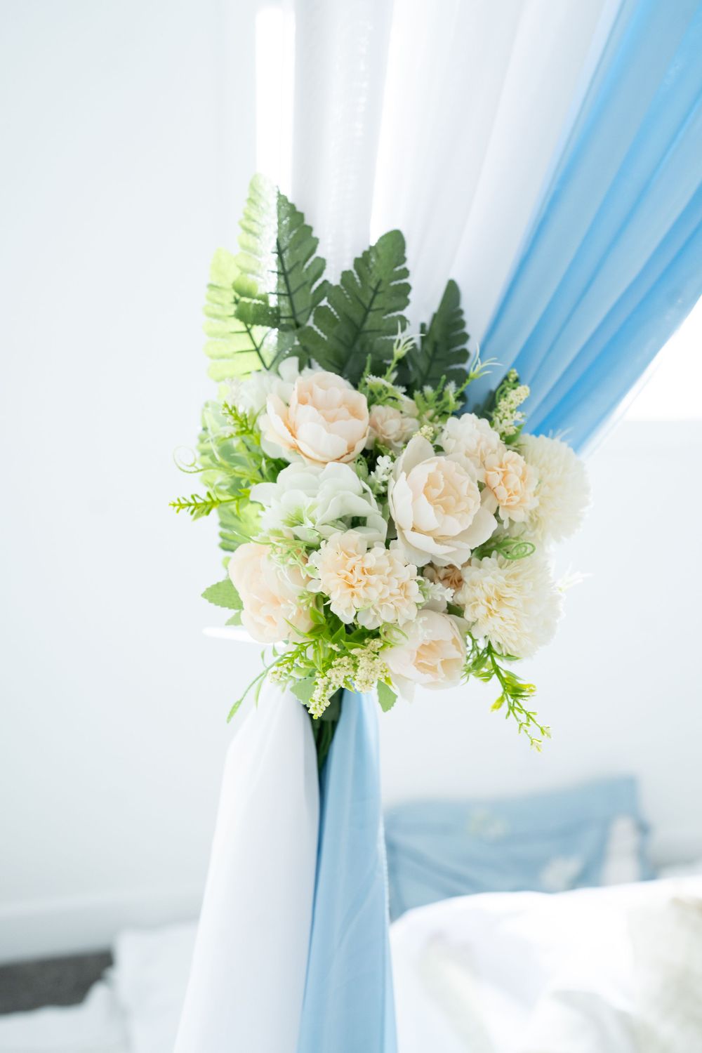 White floral arrangement with green leaves tied to blue and white draped wedding canopy post.