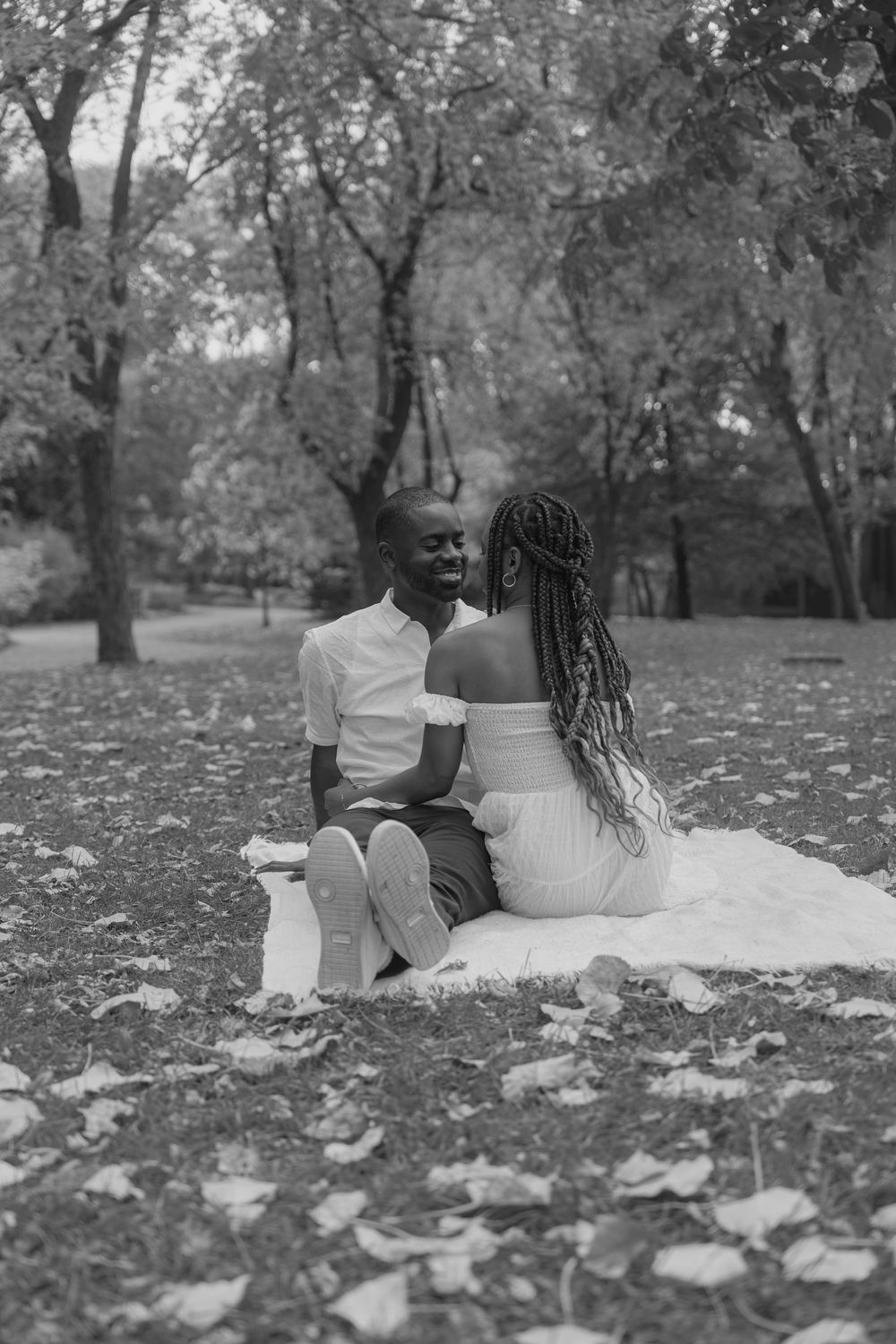 A couple sits on a white blanket surrounded by fallen autumn leaves in a park setting.