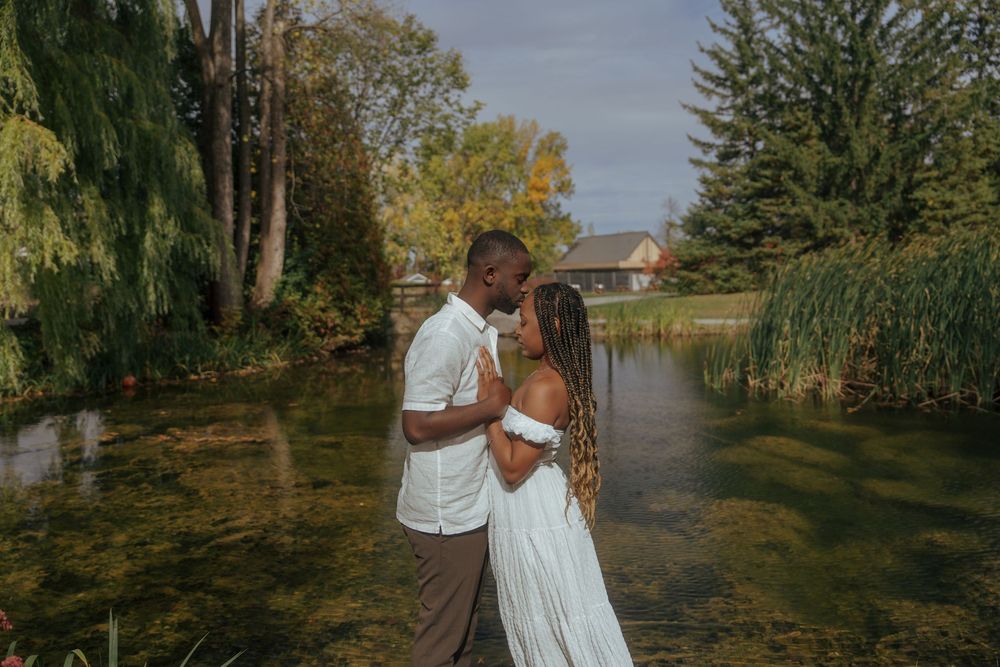 A couple shares a tender moment by a waterfront with willow trees in the background.