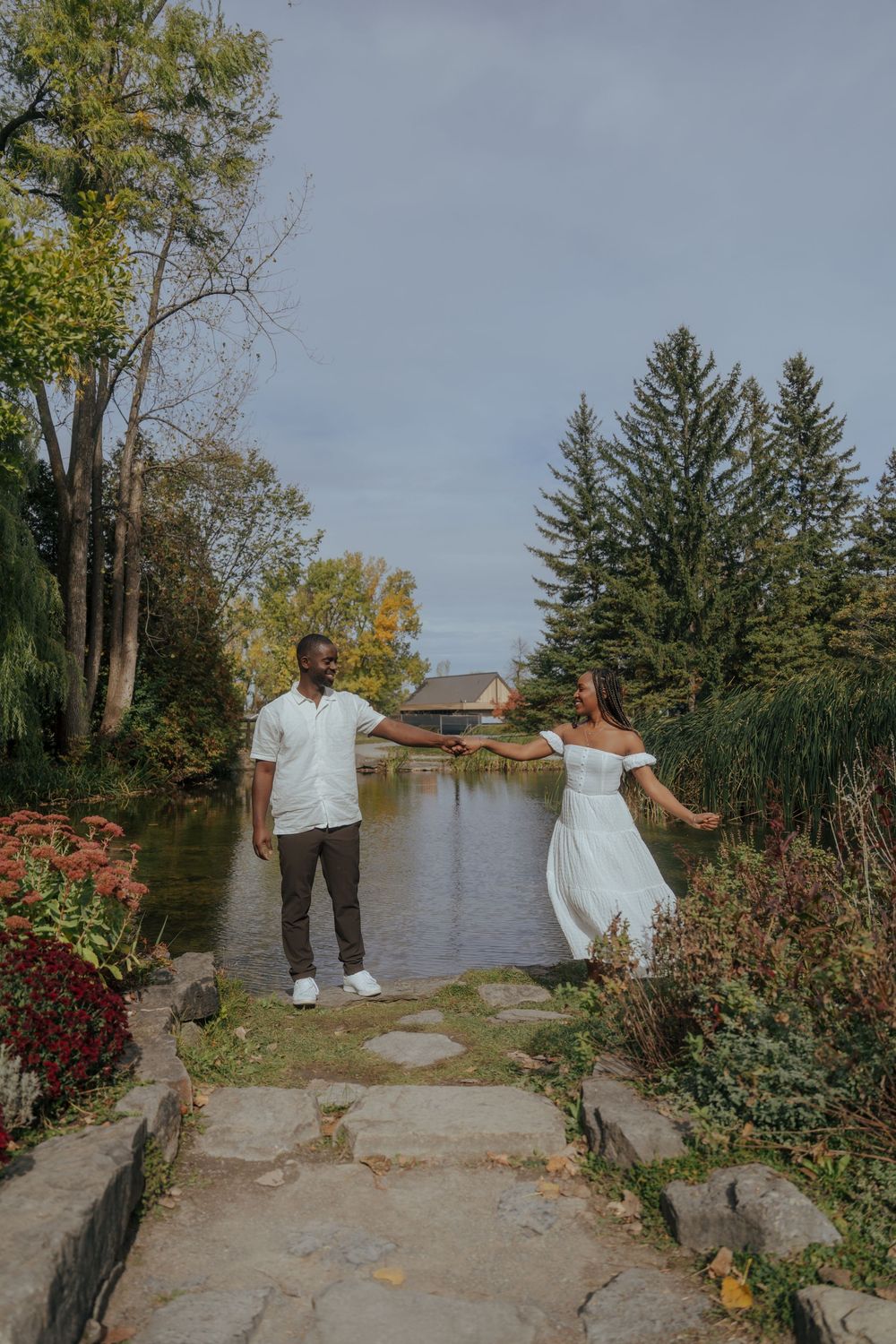 A couple stands on stone steps by a tranquil pond surrounded by autumn foliage and evergreen trees on a cloudy day.