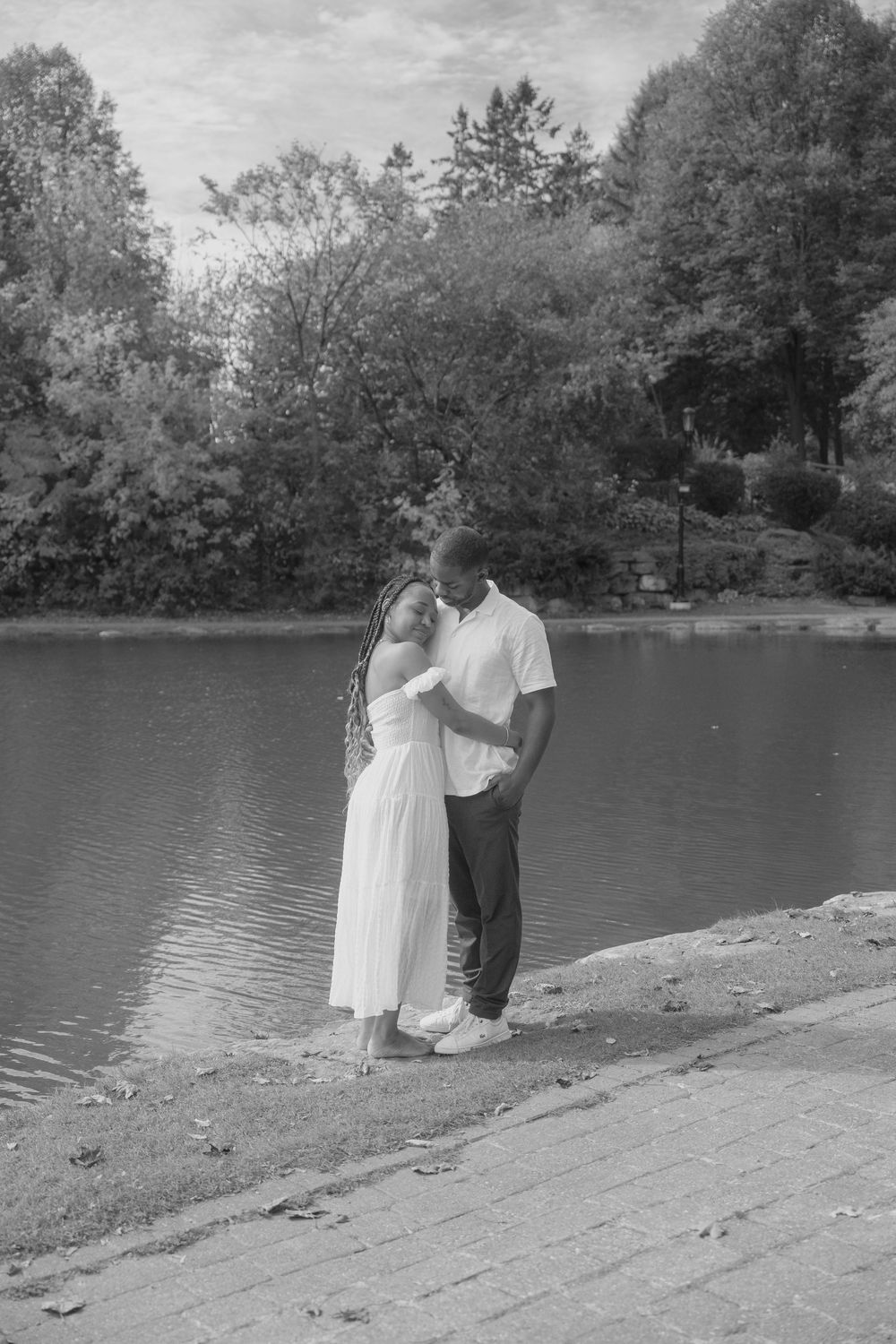A romantic black and white photo of a couple embracing by a serene lake surrounded by trees.