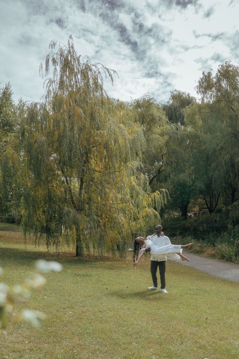 A couple embraces in a scenic park setting with willow trees and autumn foliage in the background.