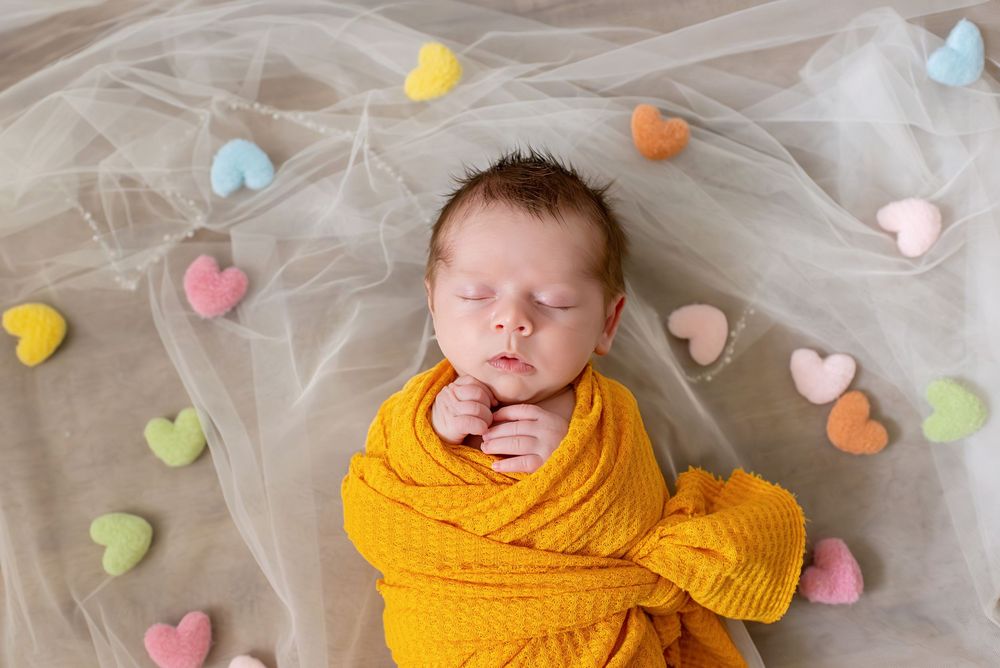 Sleeping baby wrapped in yellow knit blanket surrounded by colorful heart decorations.