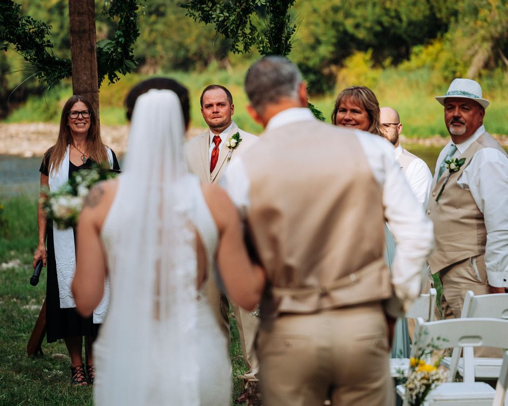 A wedding ceremony takes place outdoors with guests seated in white chairs surrounded by lush greenery and trees.
