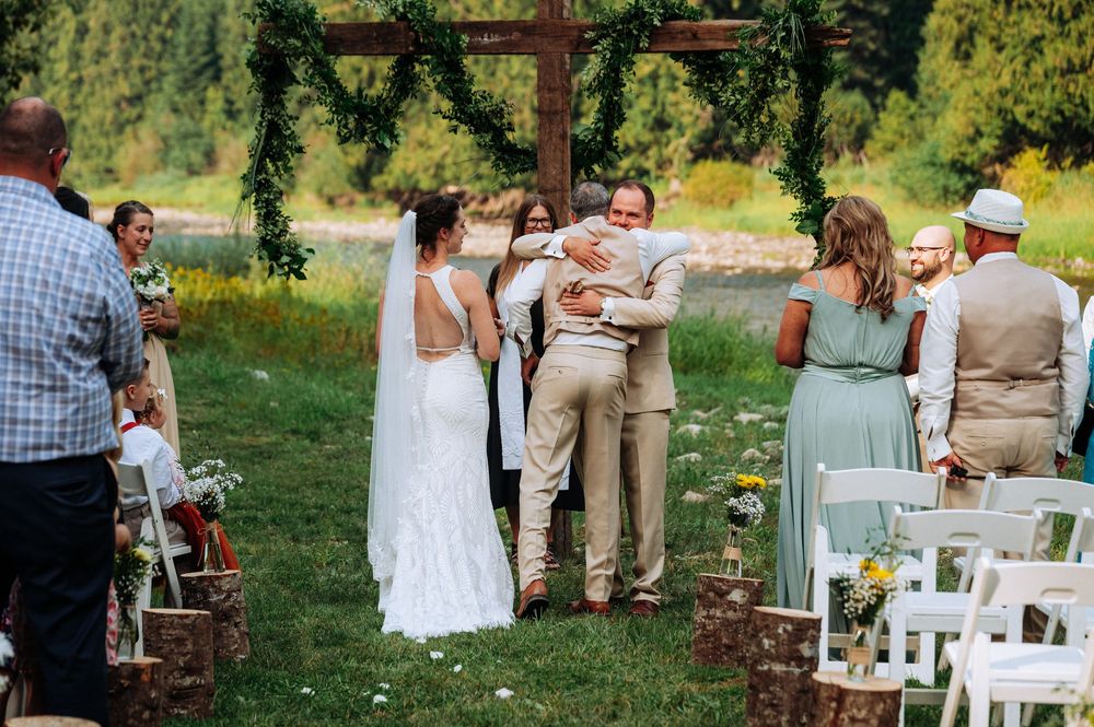 Outdoor rustic wedding ceremony takes place in front of wooden cross with greenery at scenic countryside venue.