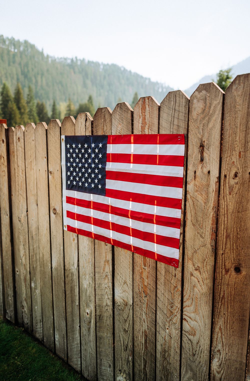 American flags mounted on weathered wooden fence panels against a mountainous backdrop create a patriotic display.