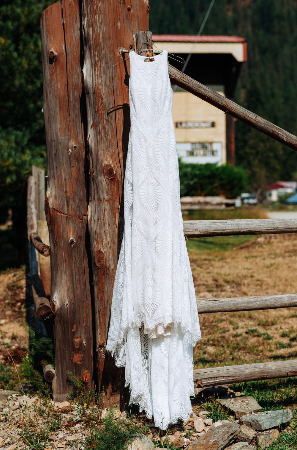 A delicate white lace wedding dress hangs on a rustic wooden fence post in an outdoor setting.