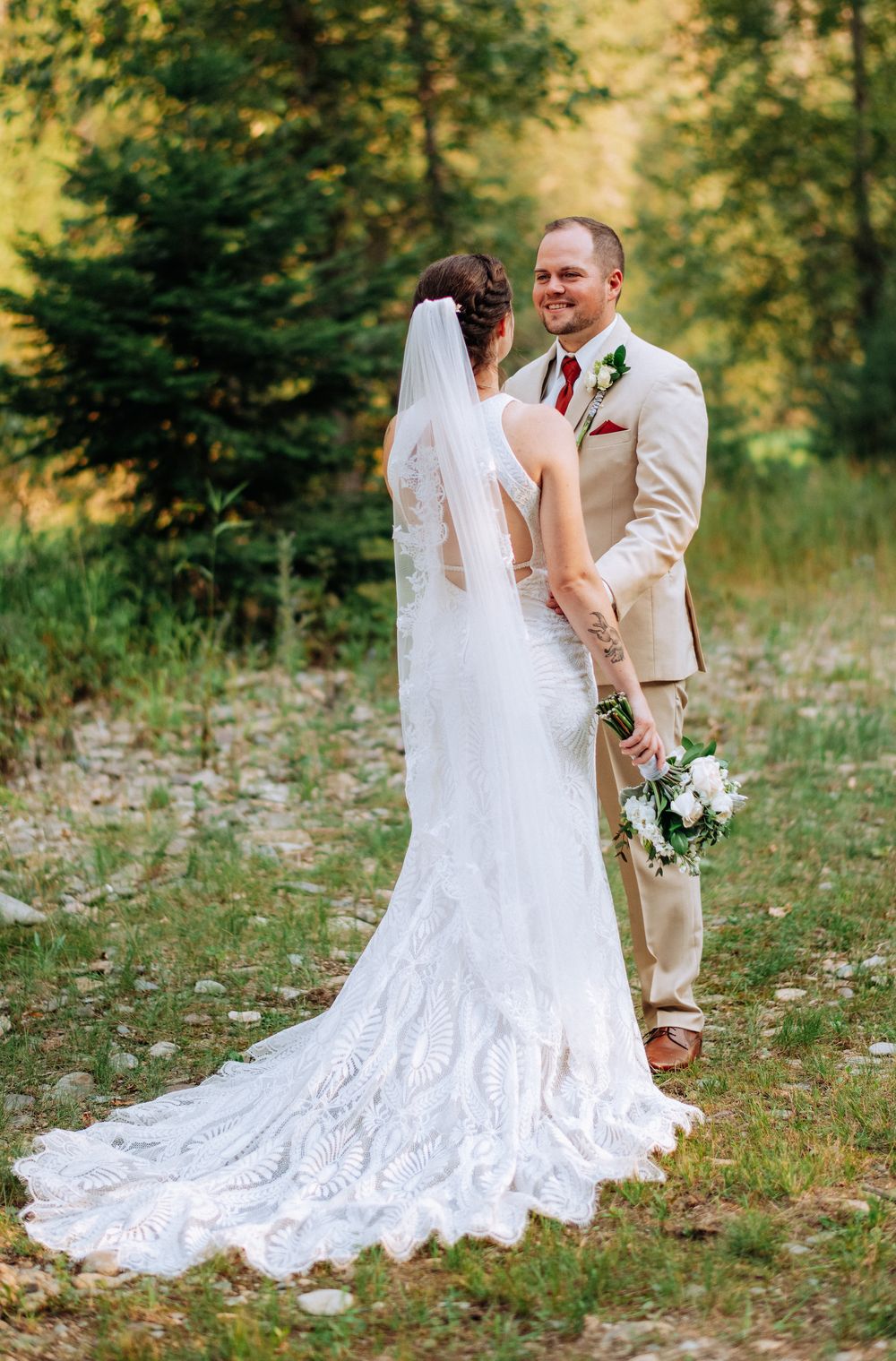 A wedding couple embraces in a romantic outdoor forest setting with sunlit trees and white lace dress details.