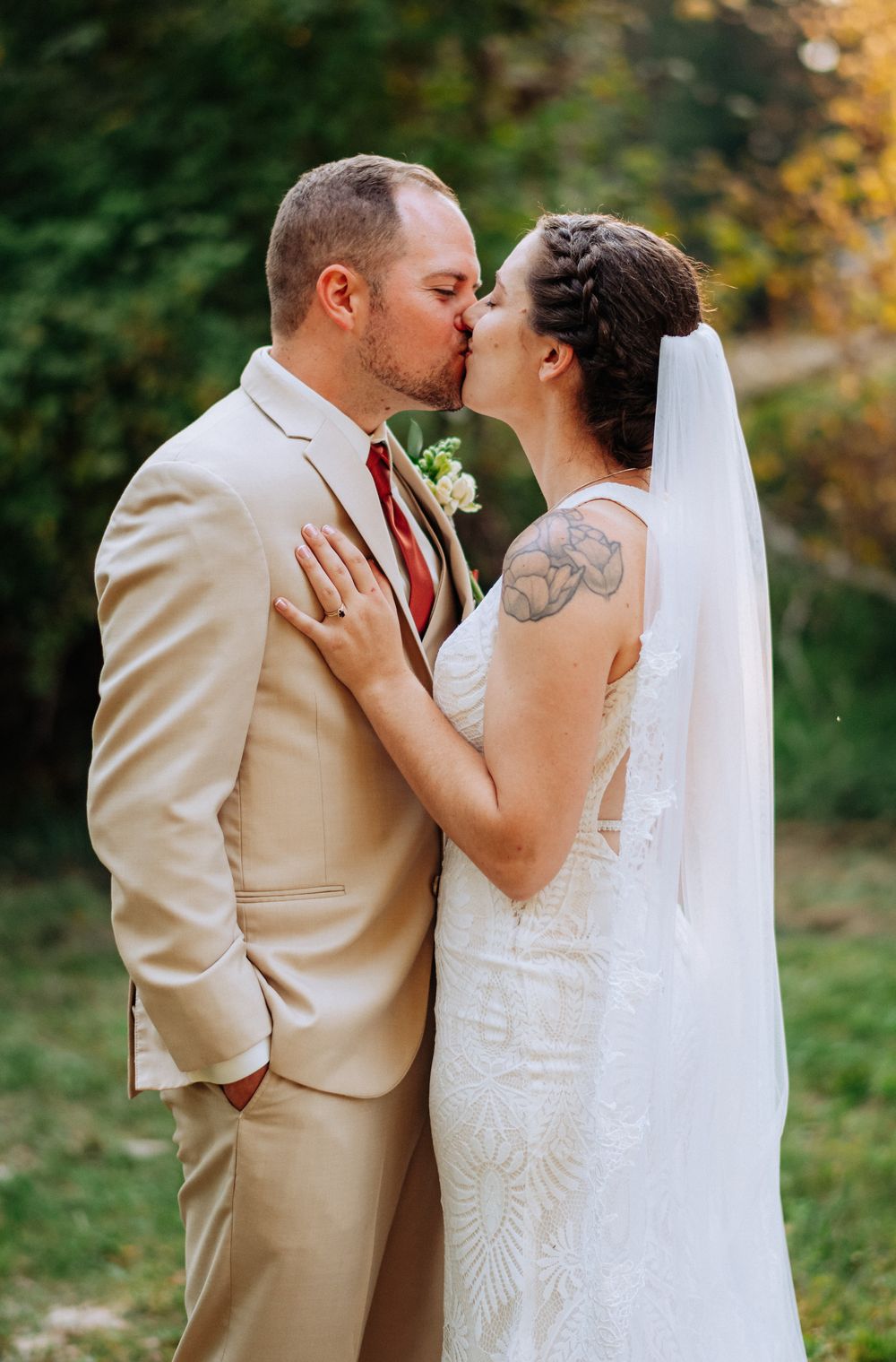 A couple shares a romantic kiss on their wedding day, with the bride in white lace and groom in tan suit outdoors.