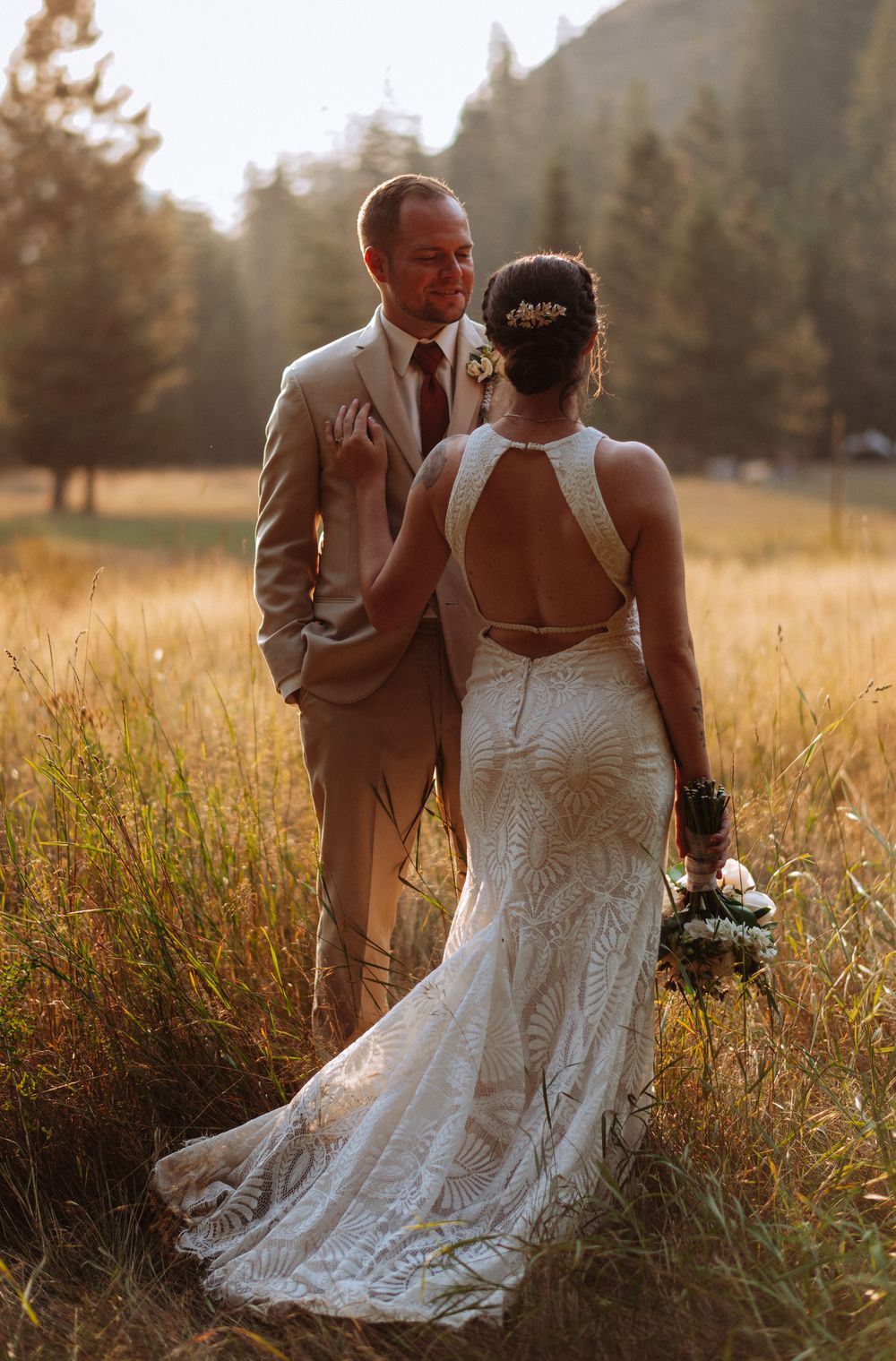 A couple in wedding attire embraces in a sunlit field of tall grass during golden hour.