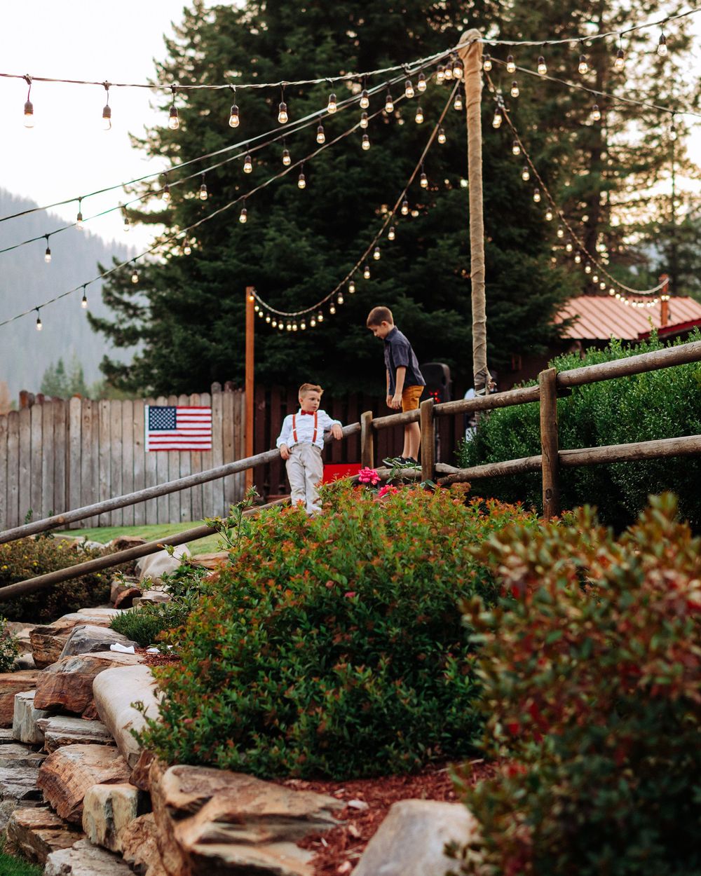 String lights illuminate a rustic outdoor patio with stone steps and lush greenery at sunset.