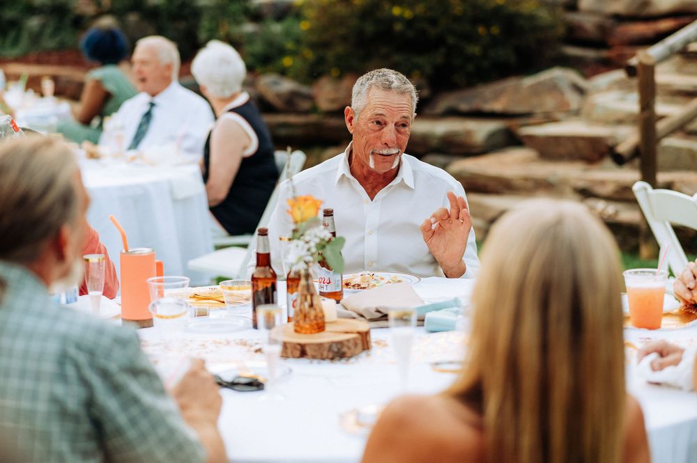 A group of people enjoying a festive outdoor meal at a long white-clothed table in a garden setting.