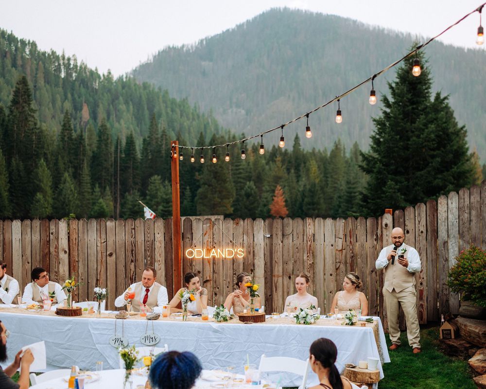 String lights illuminate an outdoor wedding reception with mountain views and wooden fencing at dusk.