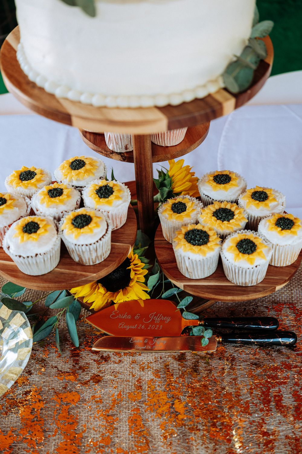 Rustic wedding dessert display featuring sunflower-decorated cupcakes and a white cake on wooden stands.