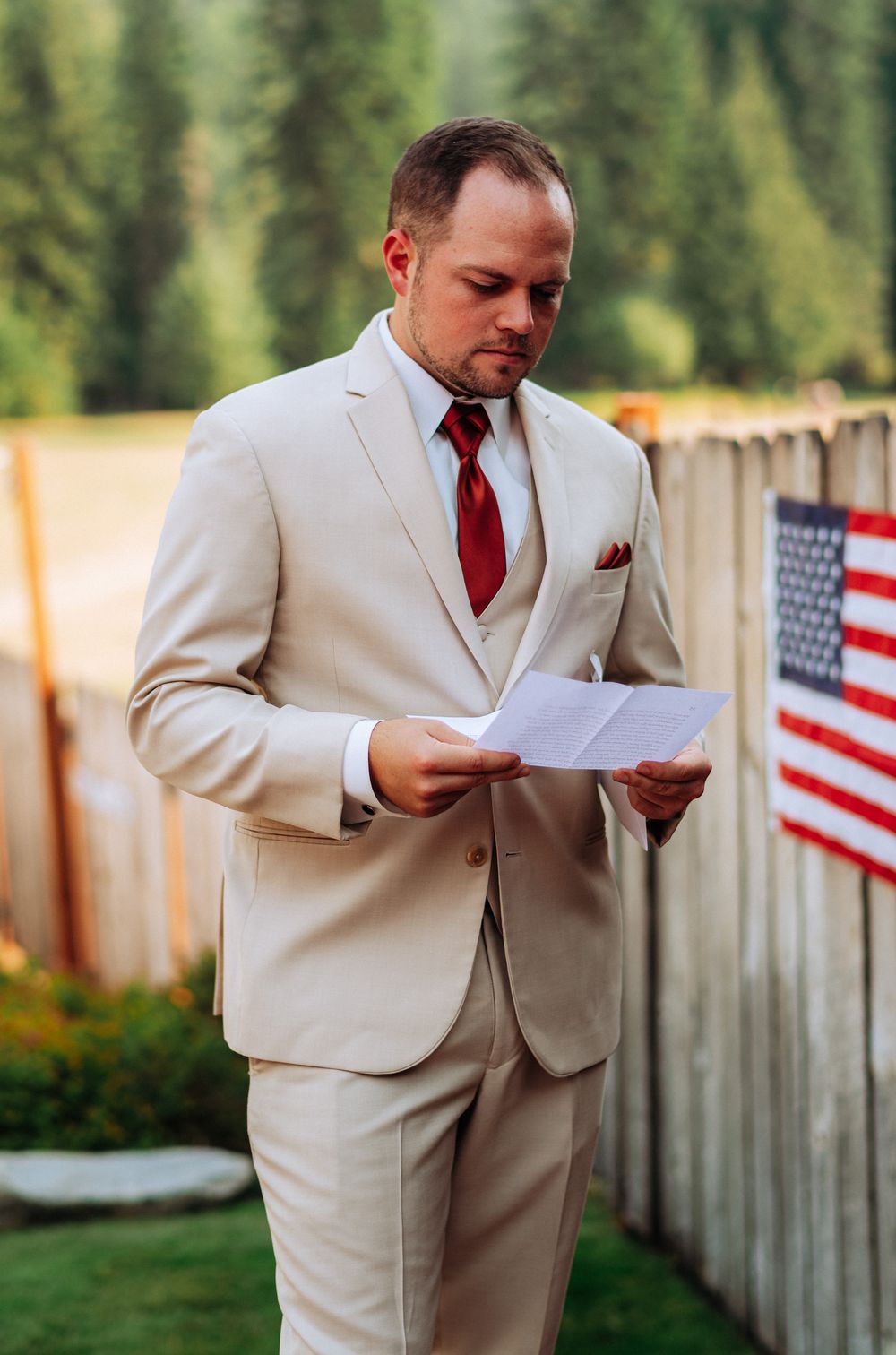 Someone in a beige suit and red tie reads a letter outdoors with American flags visible in the background.