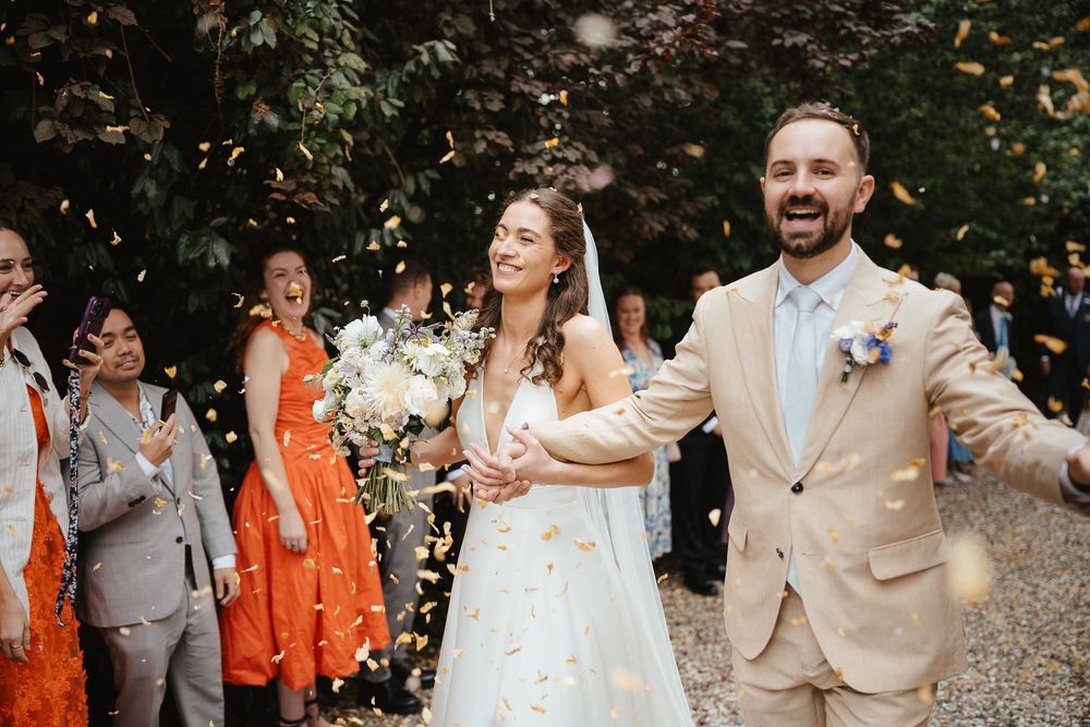 Wedding party celebrates with confetti toss in outdoor garden setting with orange and neutral color scheme.