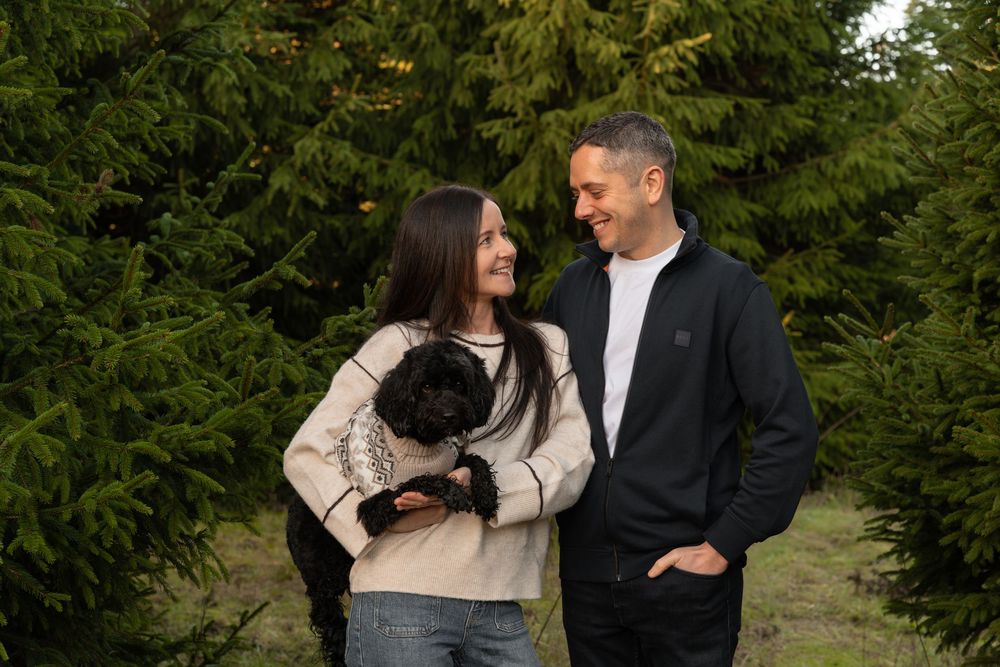 A couple stands together outdoors in front of evergreen trees while holding a small black pug.