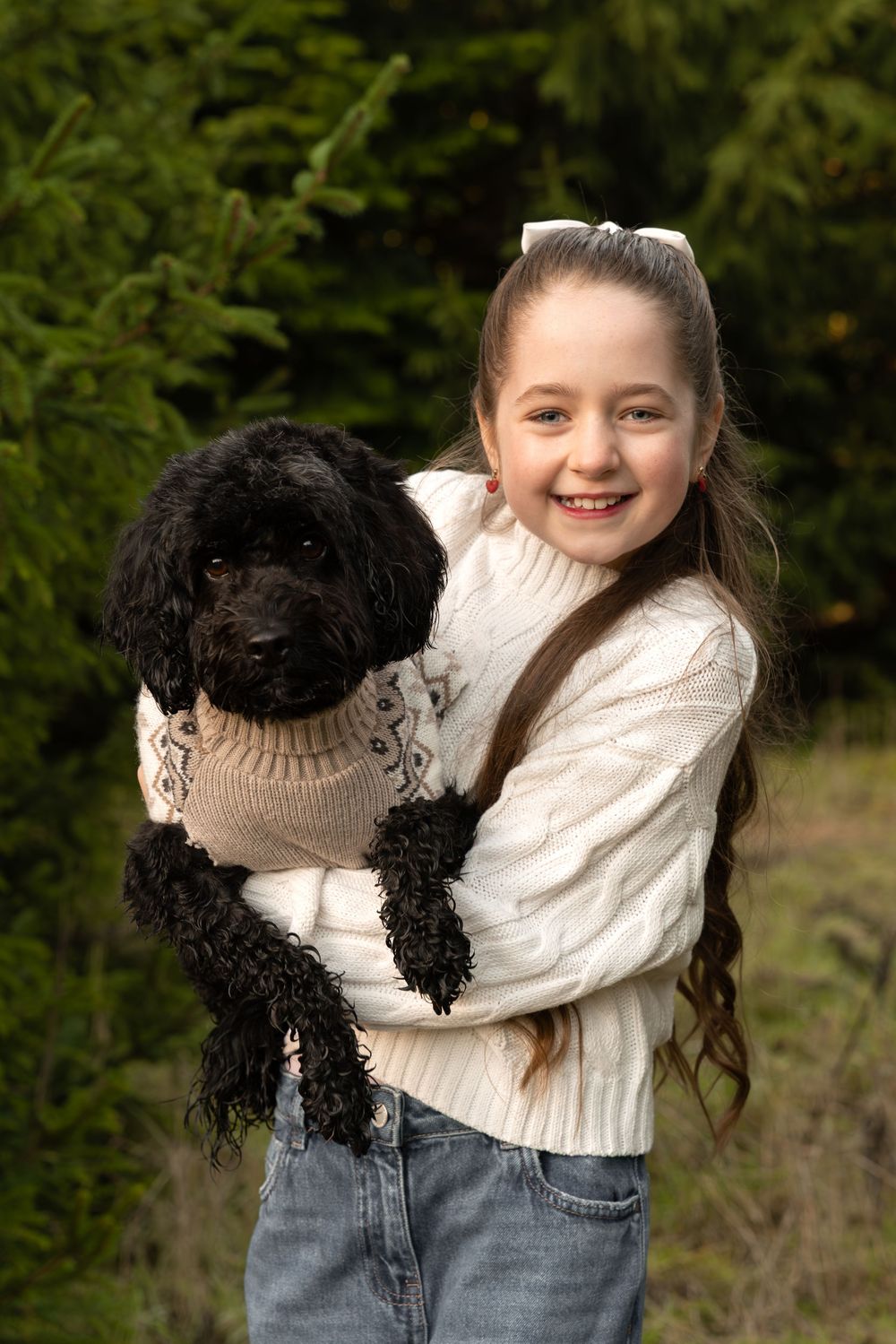 A black poodle wearing a beige sweater is held by someone in a white knit sweater and jeans outdoors.