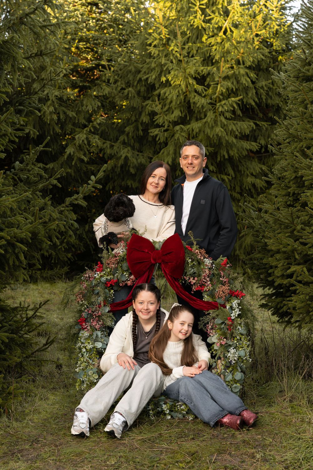 A family group poses together outdoors against evergreen trees and red berries in a winter photo session.
