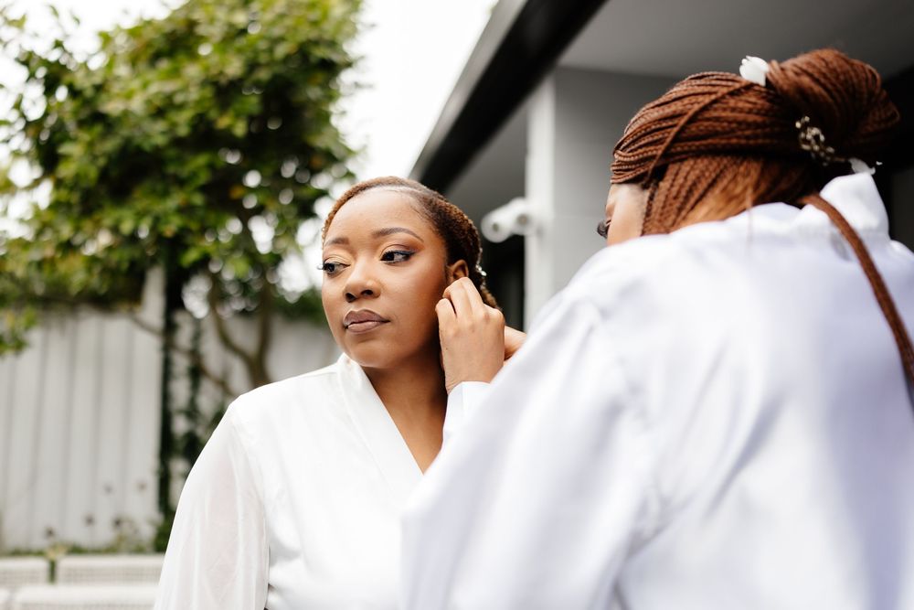 A person in white clothing puts on earrings while looking in a mirror outdoors.