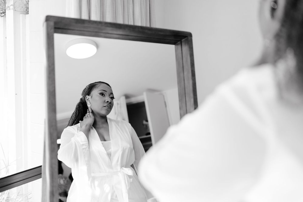 A bride getting ready in a white robe while looking in a mirror during wedding preparations.