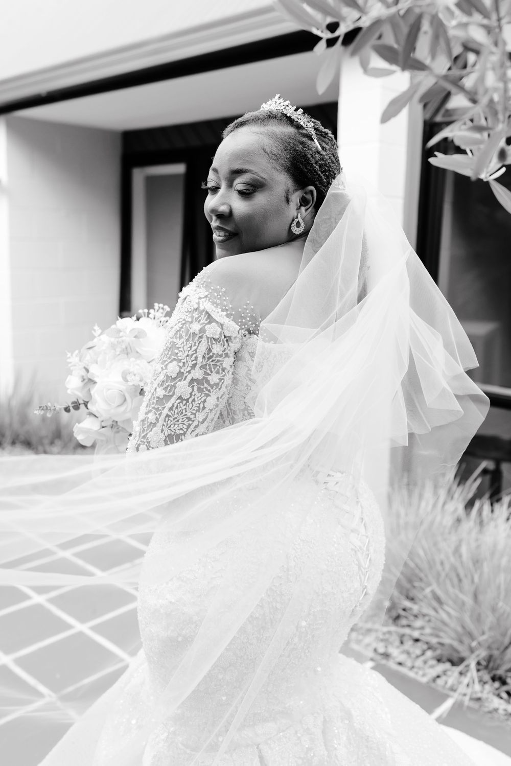 A bride in an elegant lace wedding dress with a flowing veil poses during a black and white portrait session.
