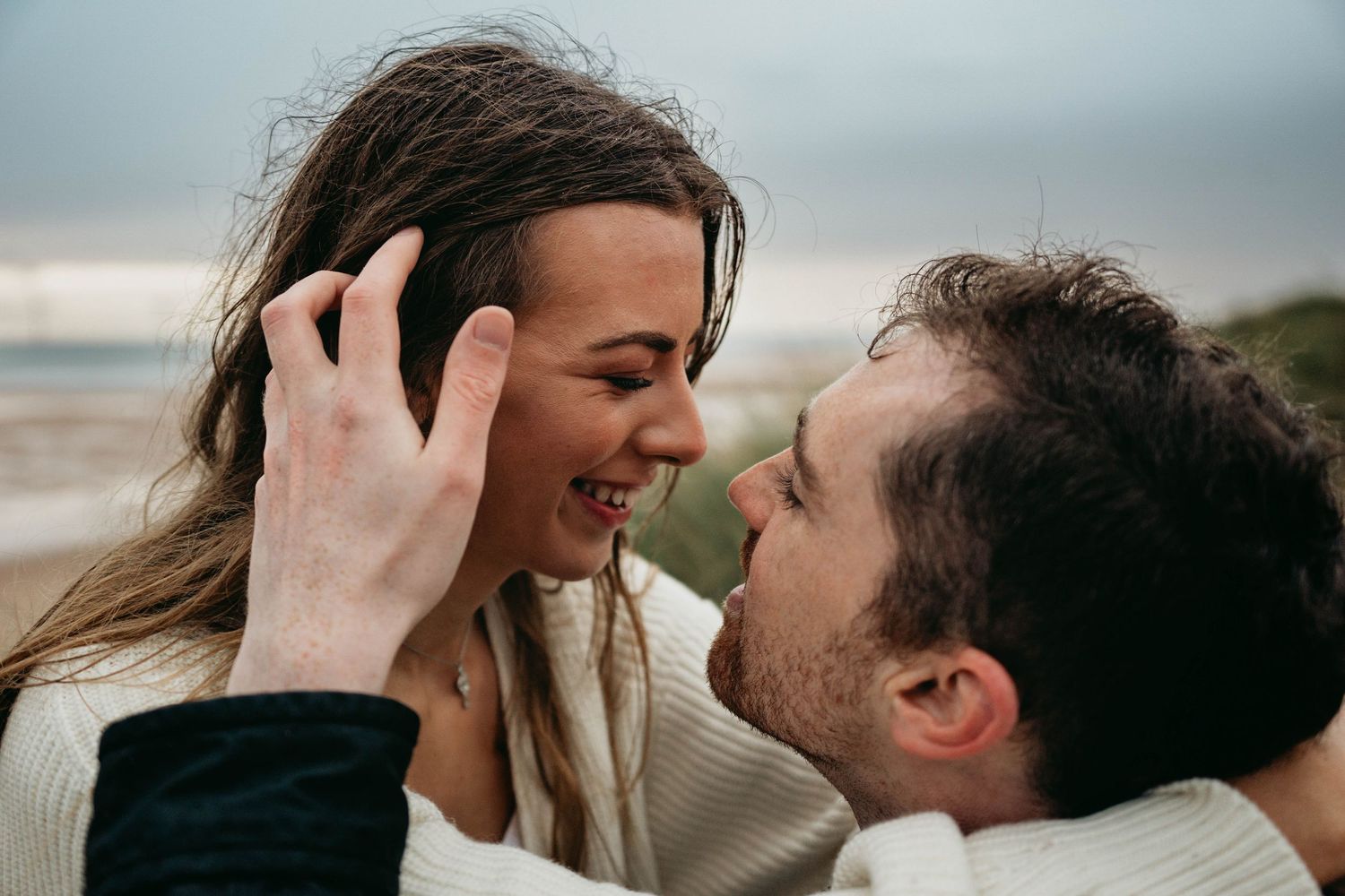 Man puts hair behind woman's ear in romantic beach engagement session.
