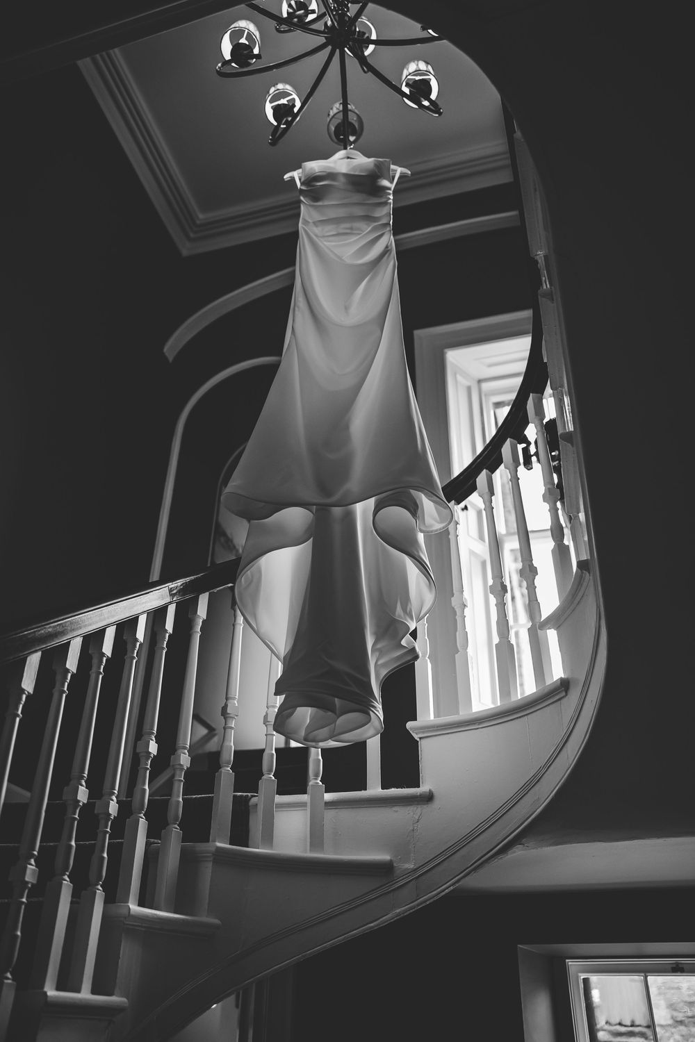 White wedding dress hangs elegantly on a curved staircase with natural light streaming through windows. Bellingham Castle Conlon Photo Film