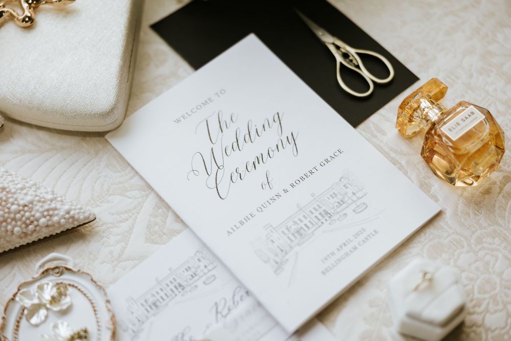 Wedding invitations and perfume arranged on a white textured surface with elegant calligraphy details.  Bellingham Castle Conlon Photo Film